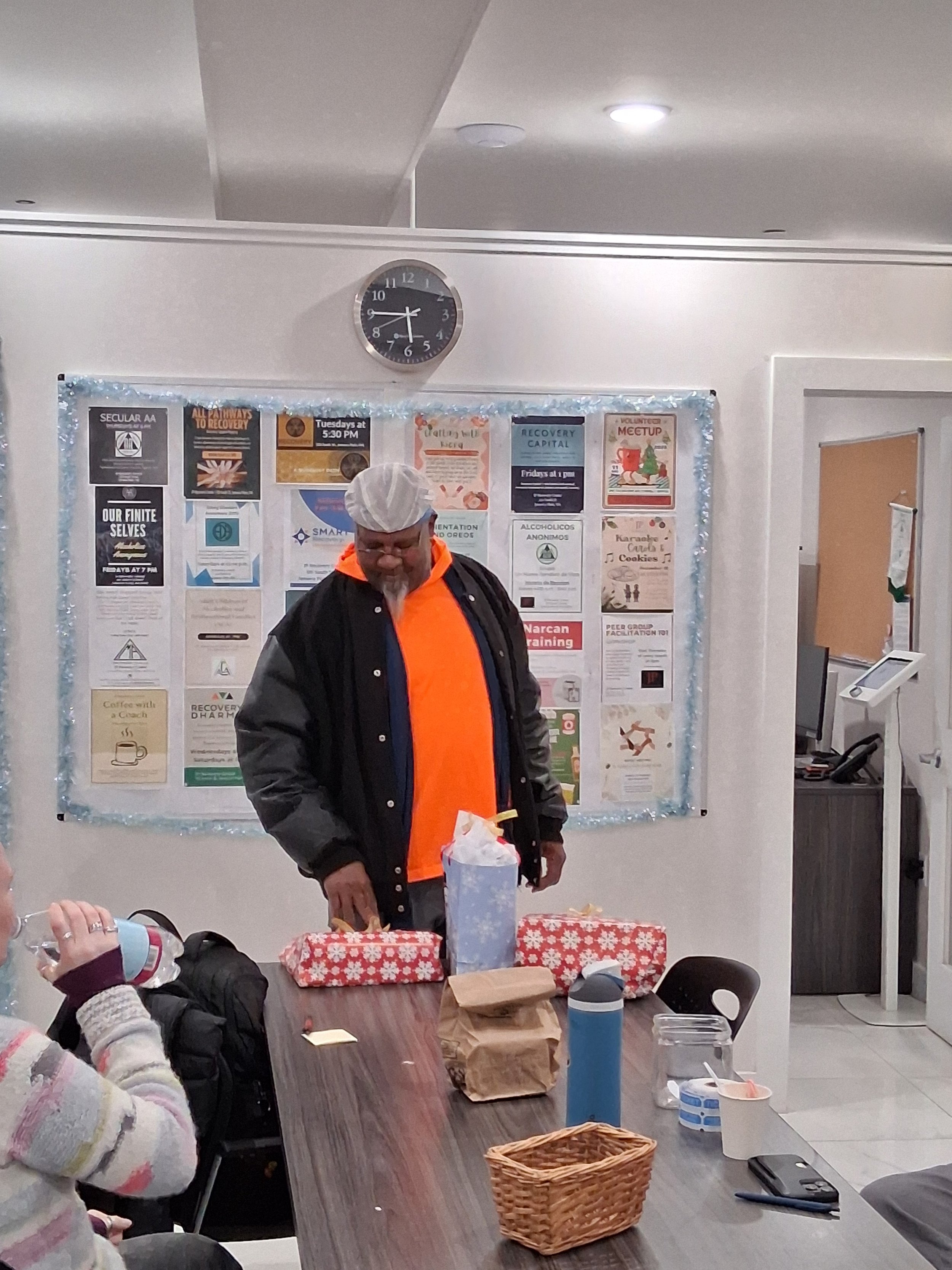 A man wearing a white hairnet and black jacket standing behind a table with wrapped presents, a tissue box, and a water bottle. A woman in a striped sweater is seated at the table to the left, drinking from a bottle. The background has educational po