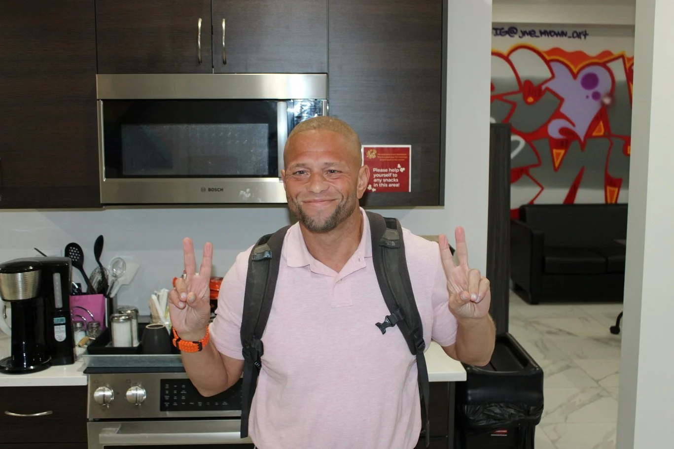 A man with a beard and short hair, wearing a pink polo shirt and a backpack, is standing in a kitchen and smiling while making peace signs with both hands.