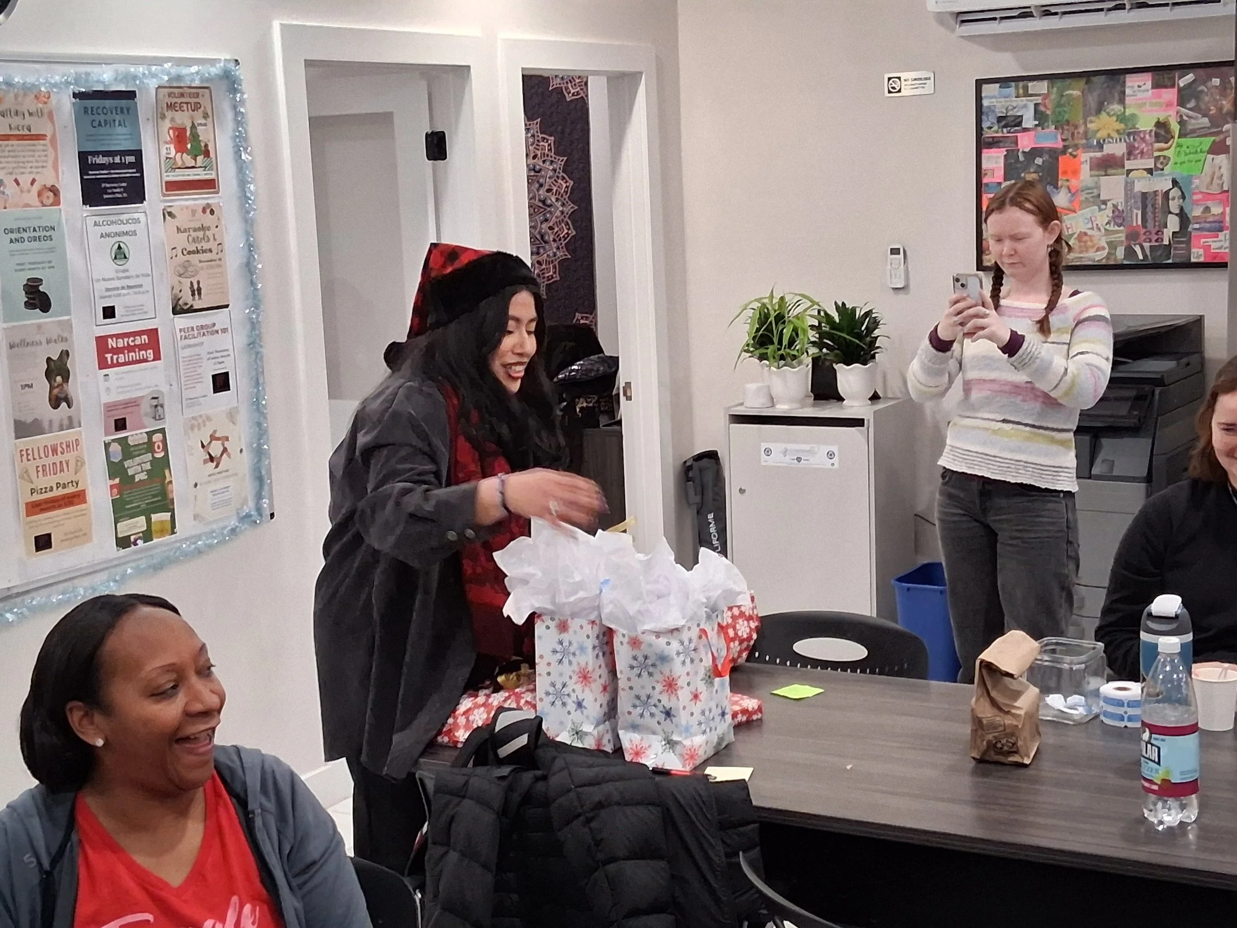 A group of women gathered around a table with Christmas-themed gift bags, one woman opening a gift, others taking photos, in a decorated room for a holiday celebration.