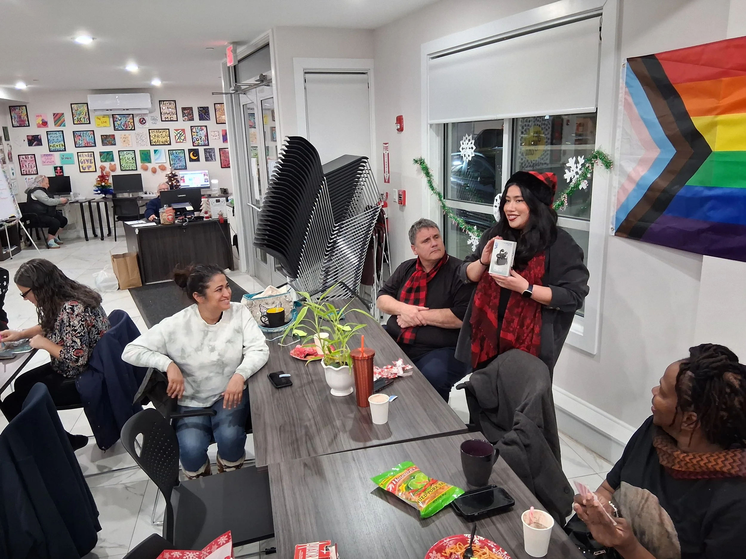 A group of people in a decorated room with holiday ornaments, one woman standing and handing out a gift or prize, others seated at a long table with snacks and drinks, in a festive setting with a rainbow pride flag on the wall.