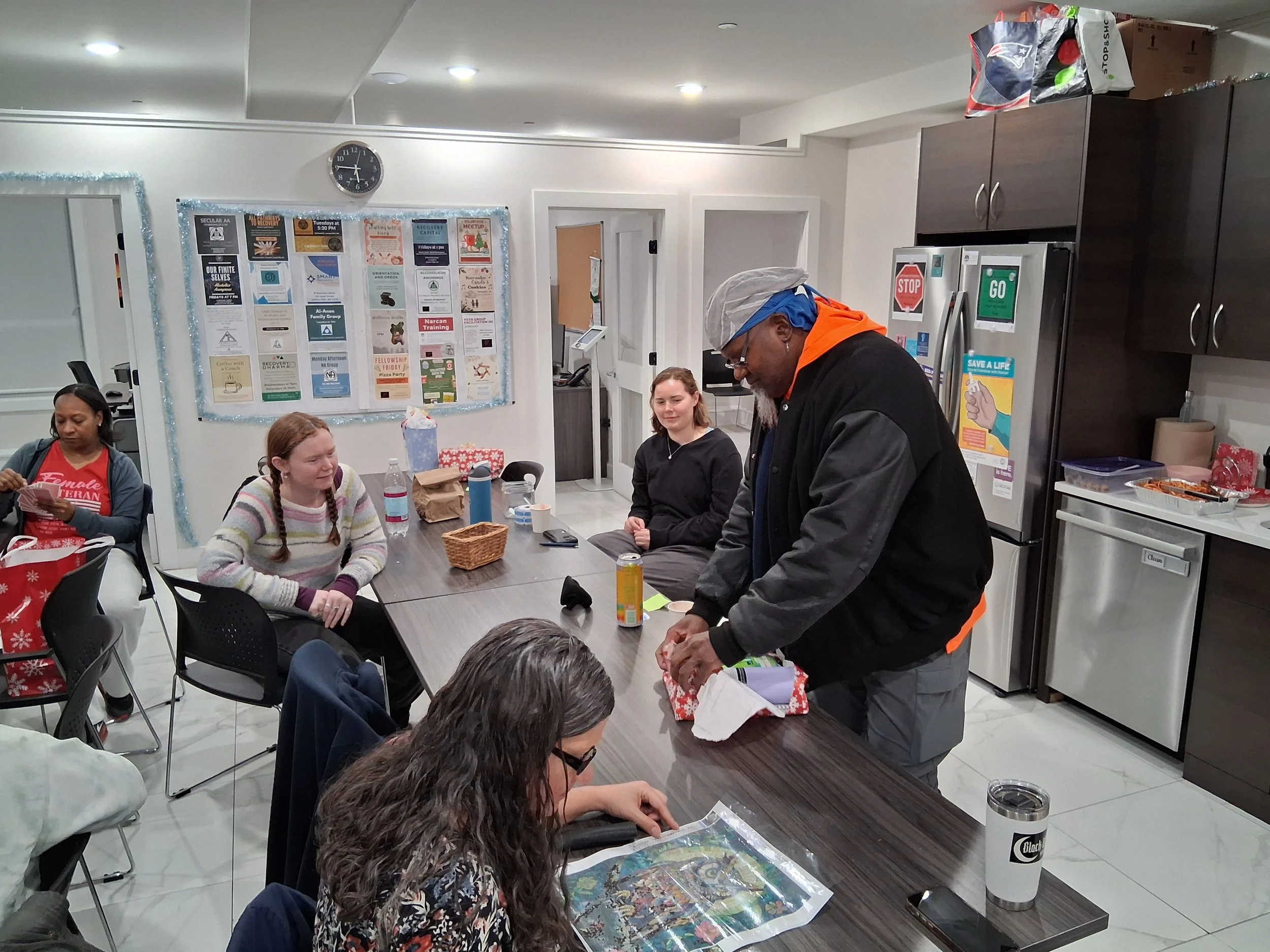 A group of people sitting around a conference table in a room with posters on the wall, some people are chatting and one man is unwrapping a gift.
