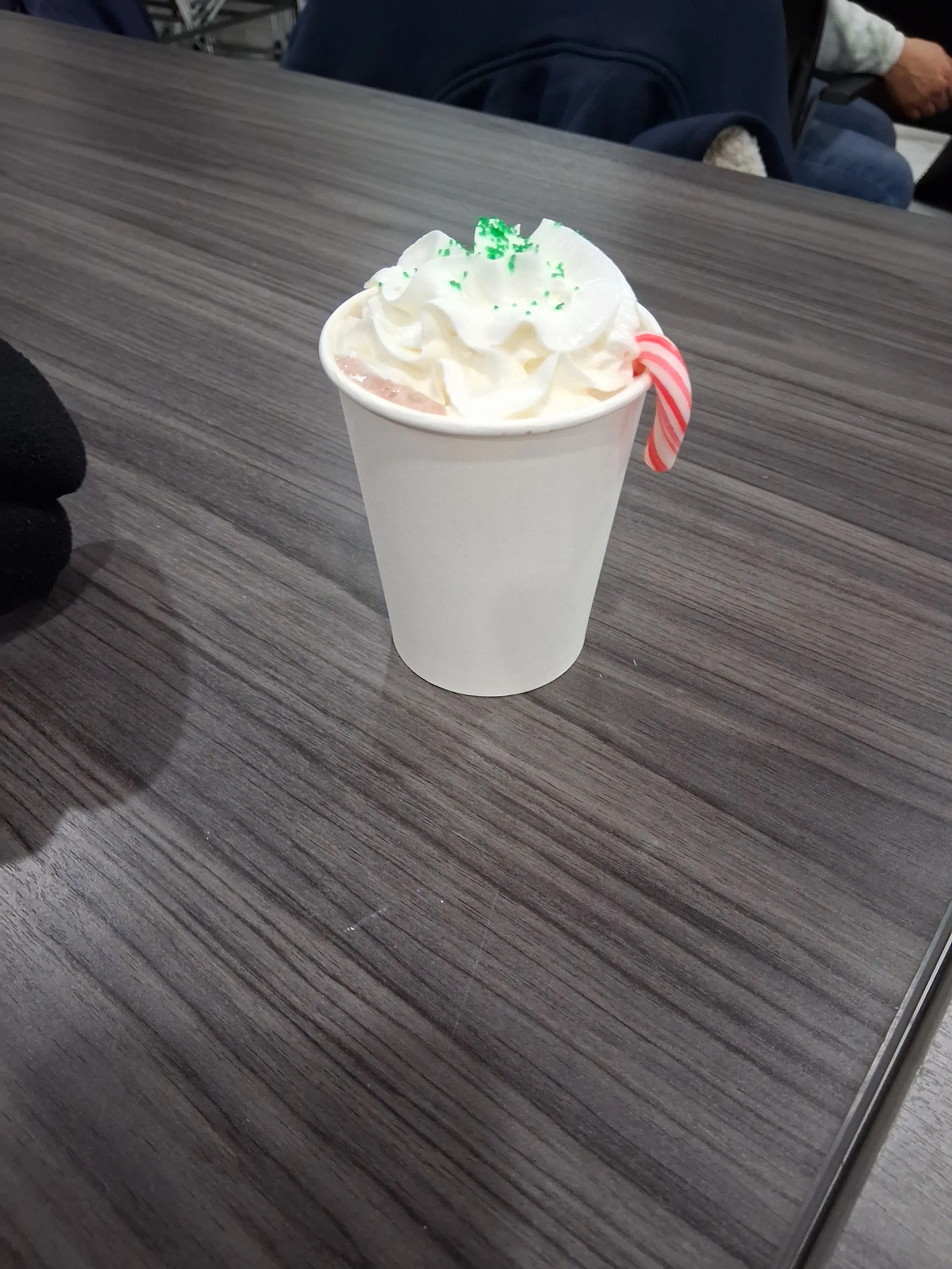 A cup of hot chocolate topped with whipped cream, green sprinkles, and a red and white candy cane on the side, placed on a dark wooden table.