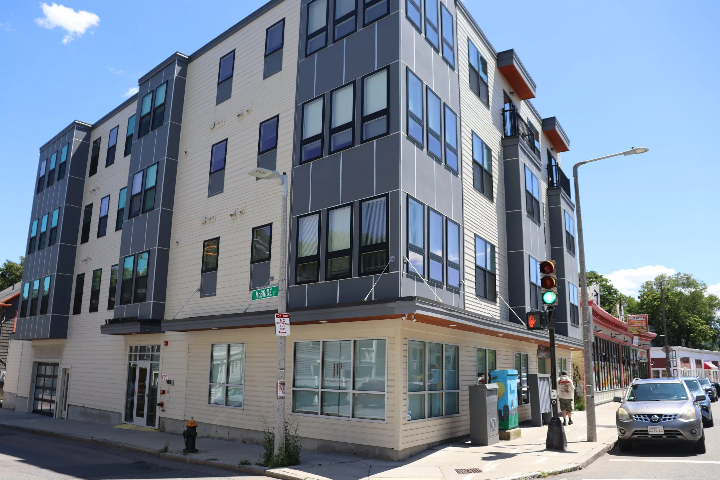 Multi-story modern building with mixed light and dark gray exterior, corner location at McBride Street, with several rectangular windows and balconies, a traffic light showing green, a sidewalk, parked cars, and a person walking.