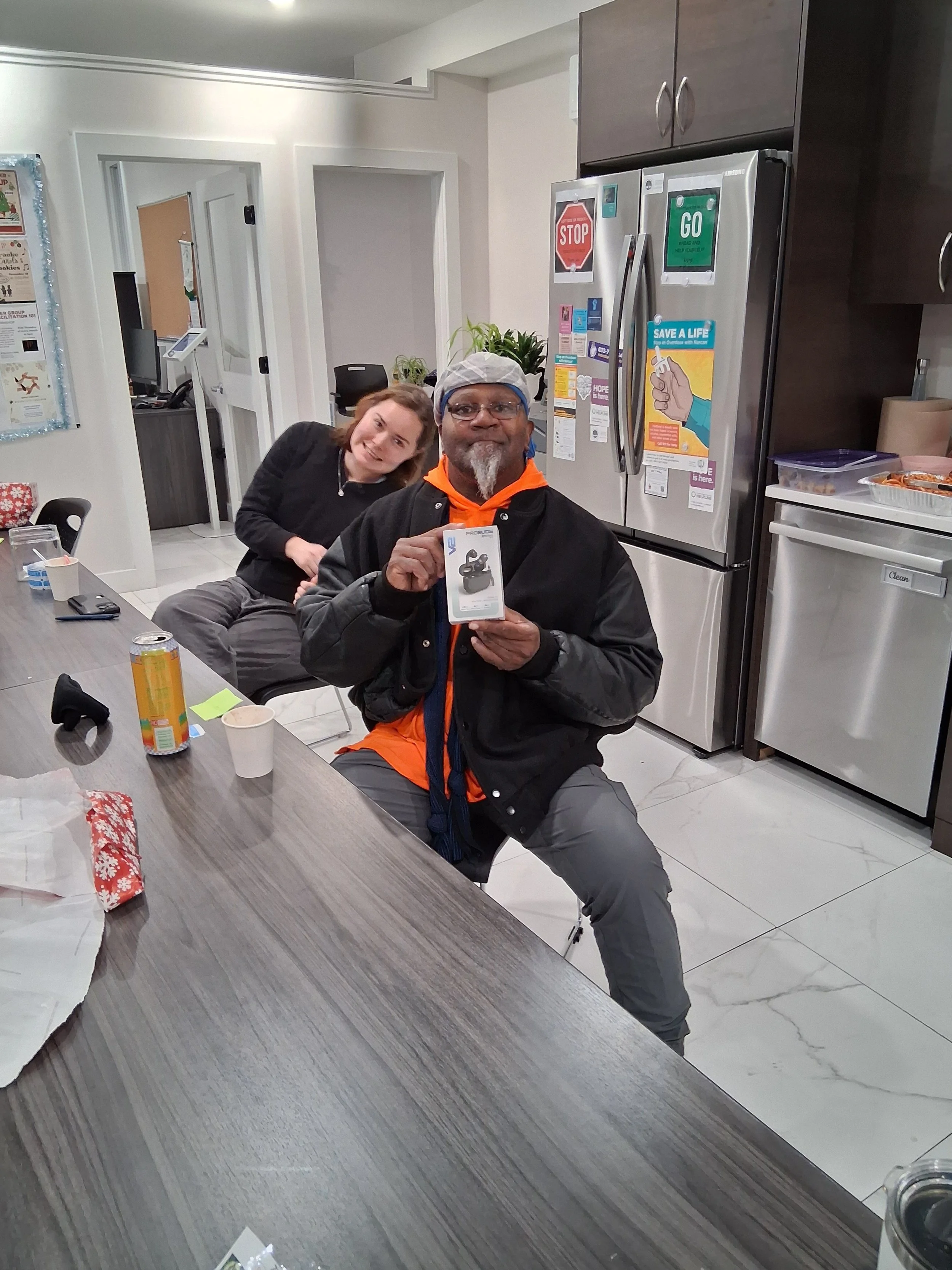 A man sitting at a table in a kitchen holding a new pair of wireless earbuds in their box, smiling. A woman behind him is leaning on the table, smiling at the camera.