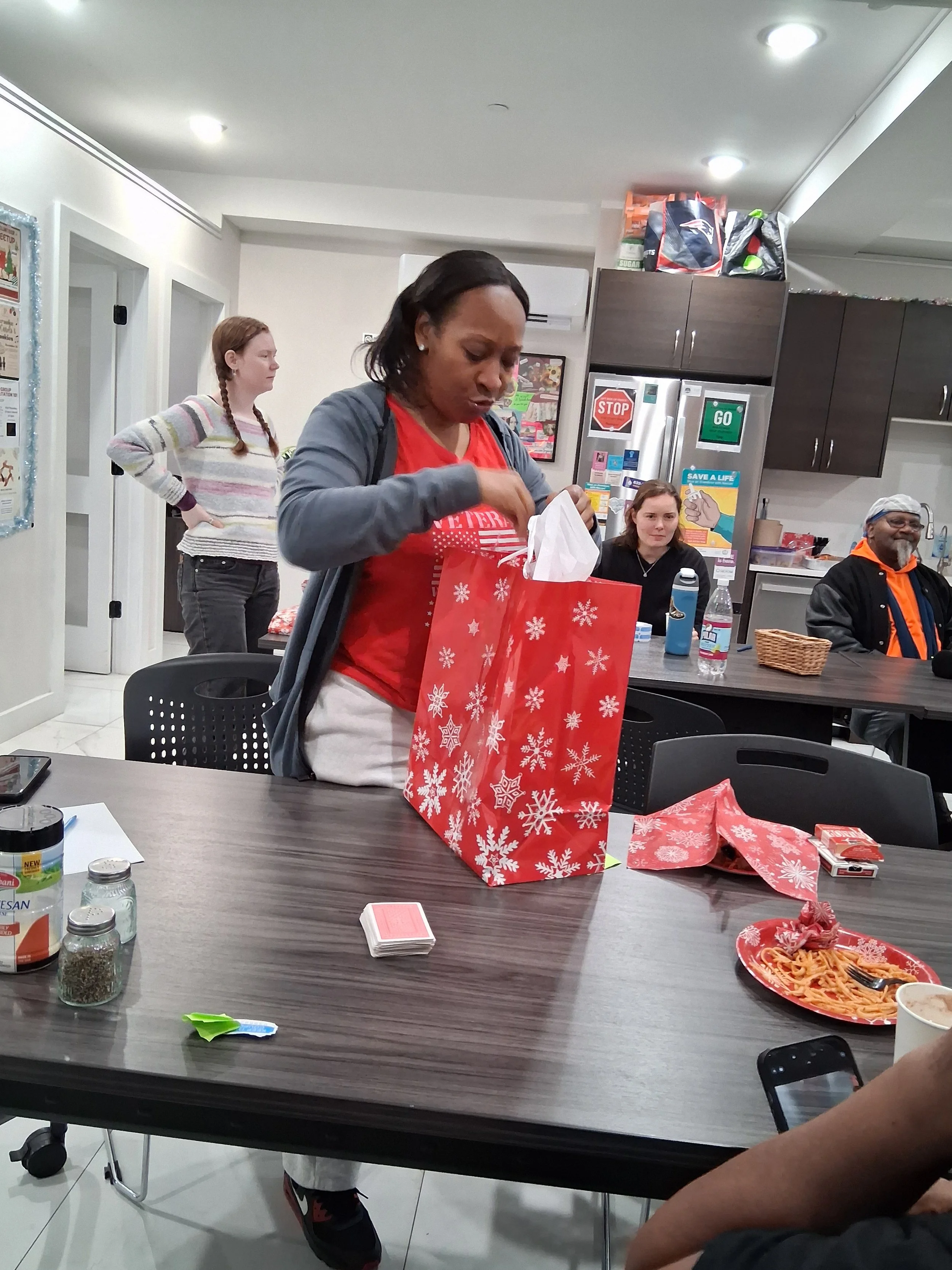 Woman opening a Christmas gift bag at a gathering, with people sitting at a table, food, and decorations in the background.