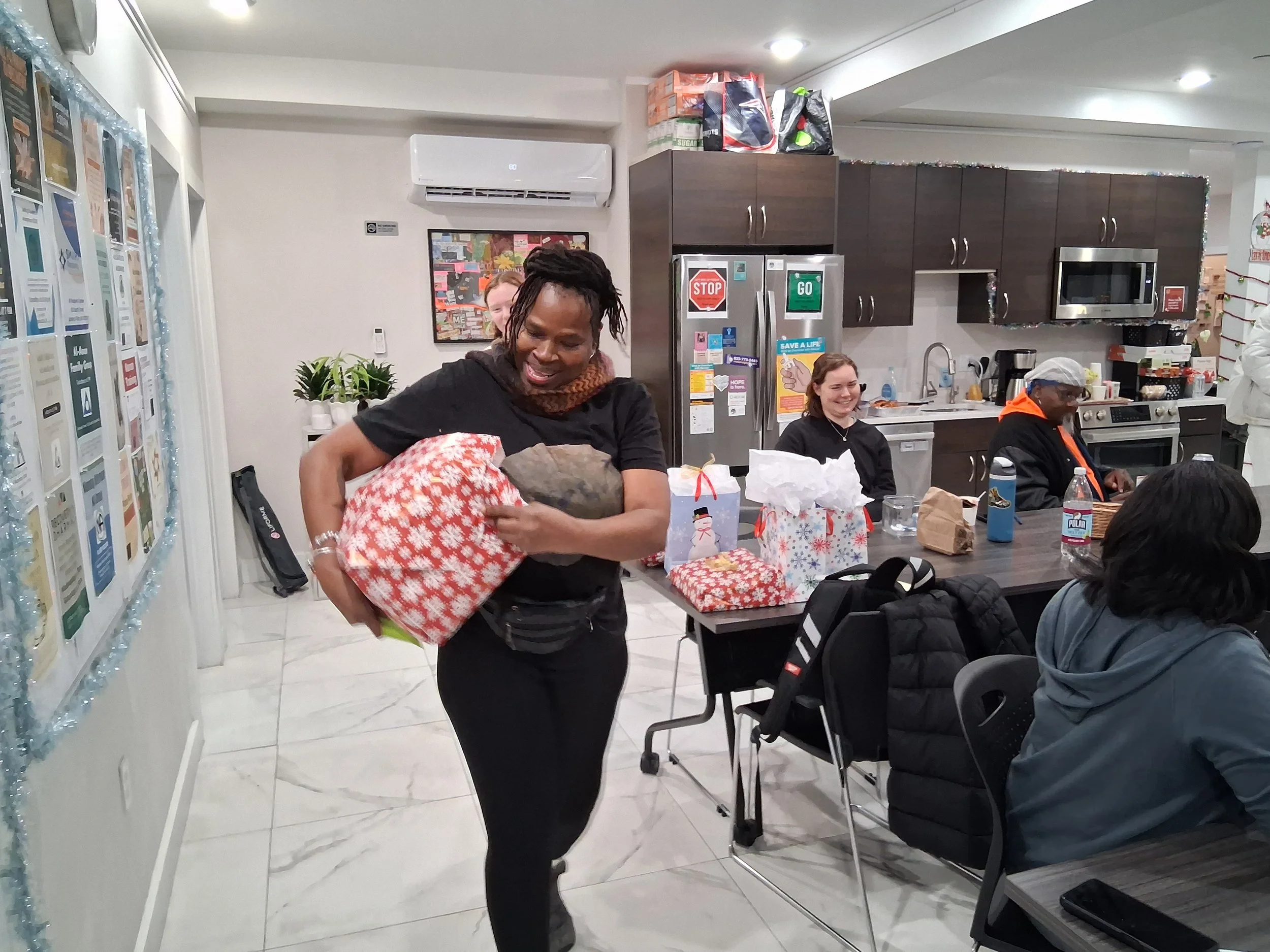 A woman smiling and holding wrapped Christmas gifts in a room with a kitchen and several people sitting at a table, surrounded by holiday decorations.