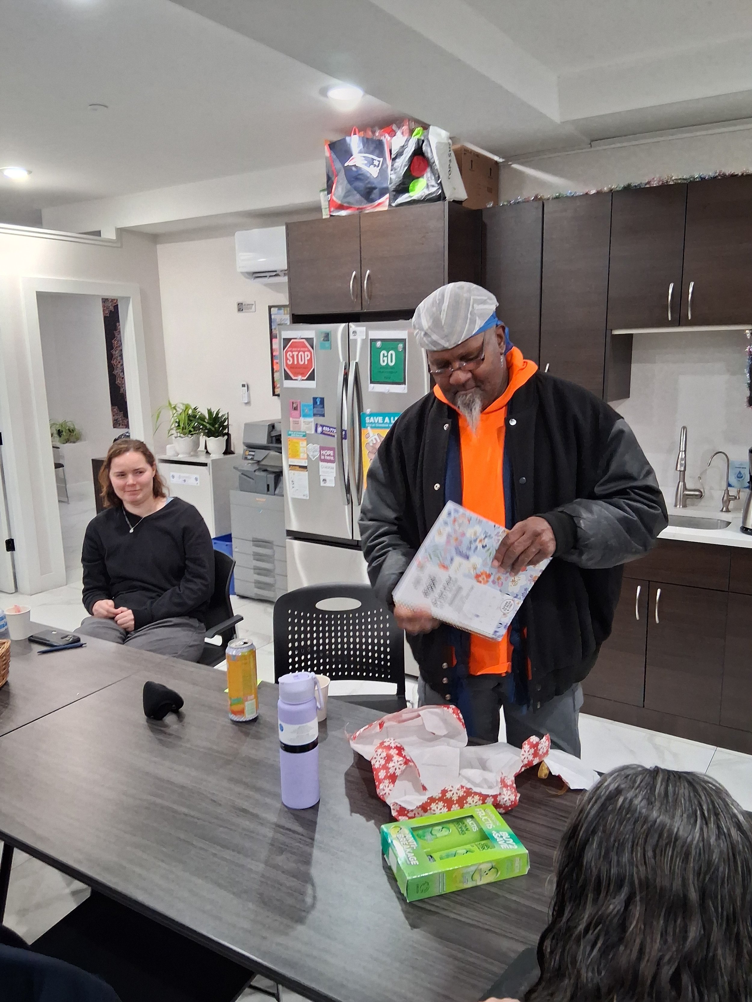 People gathered around a table in a kitchen, with a man opening a gift and a woman seated nearby.