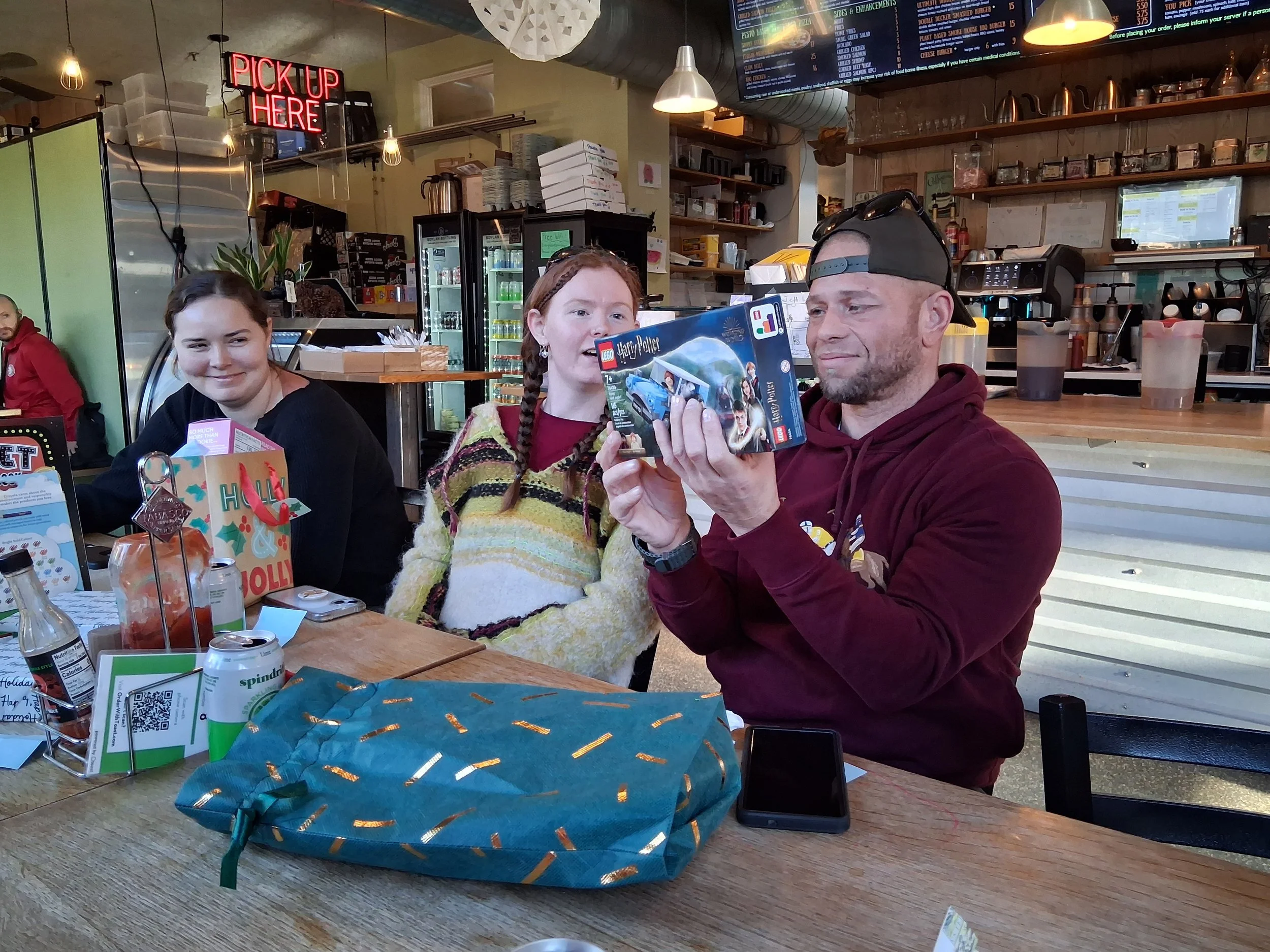 Three people sitting at a restaurant table, with the man in the red hoodie holding a Lego Harry Potter set. Two women are seated on either side, with one smiling and the other looking at the set. The restaurant has a counter in the background with a 