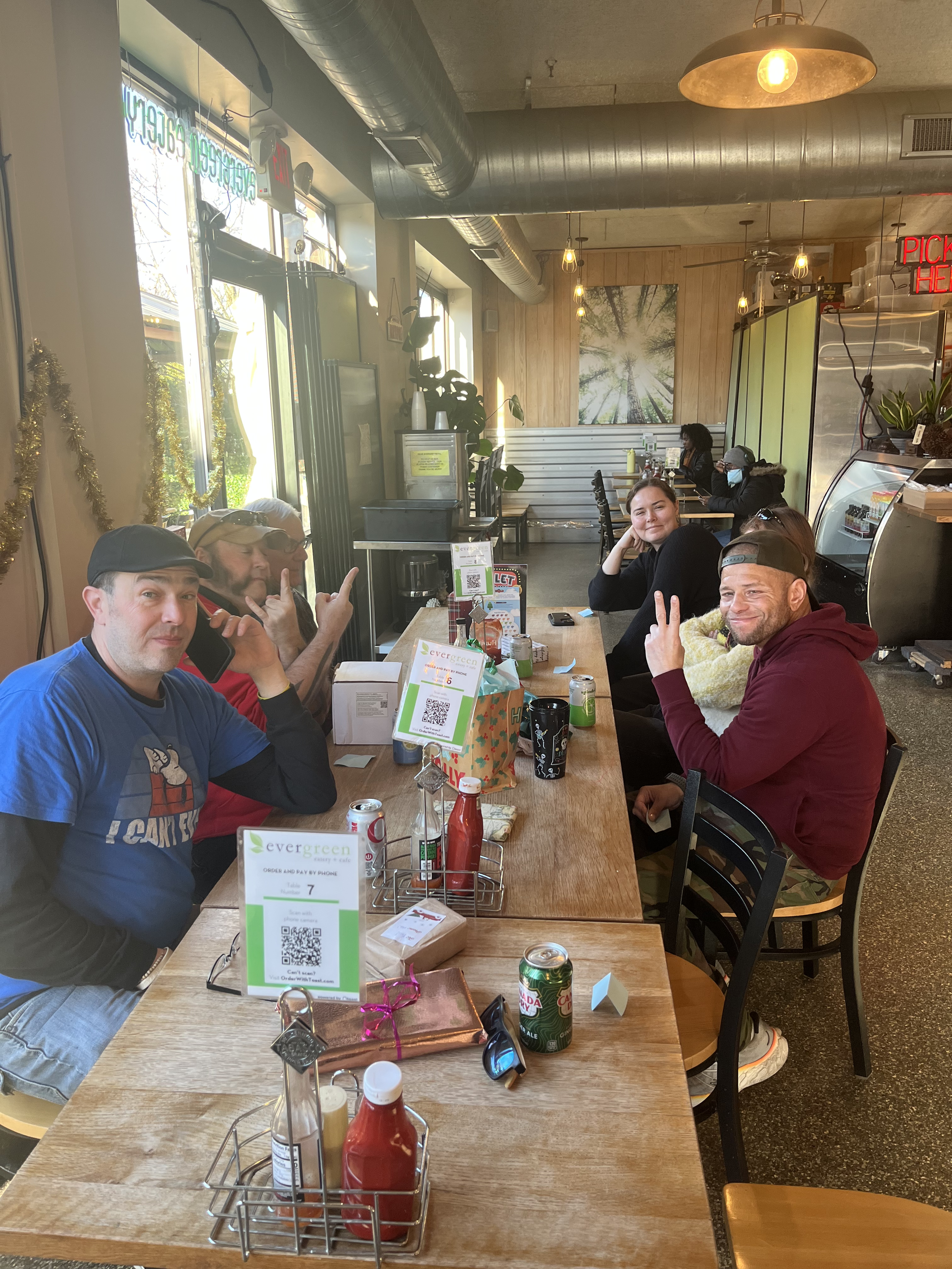 A group of six people sitting at a wooden table inside a cafe, smiling and making peace signs. The cafe has large windows, potted plants, and warm lighting.