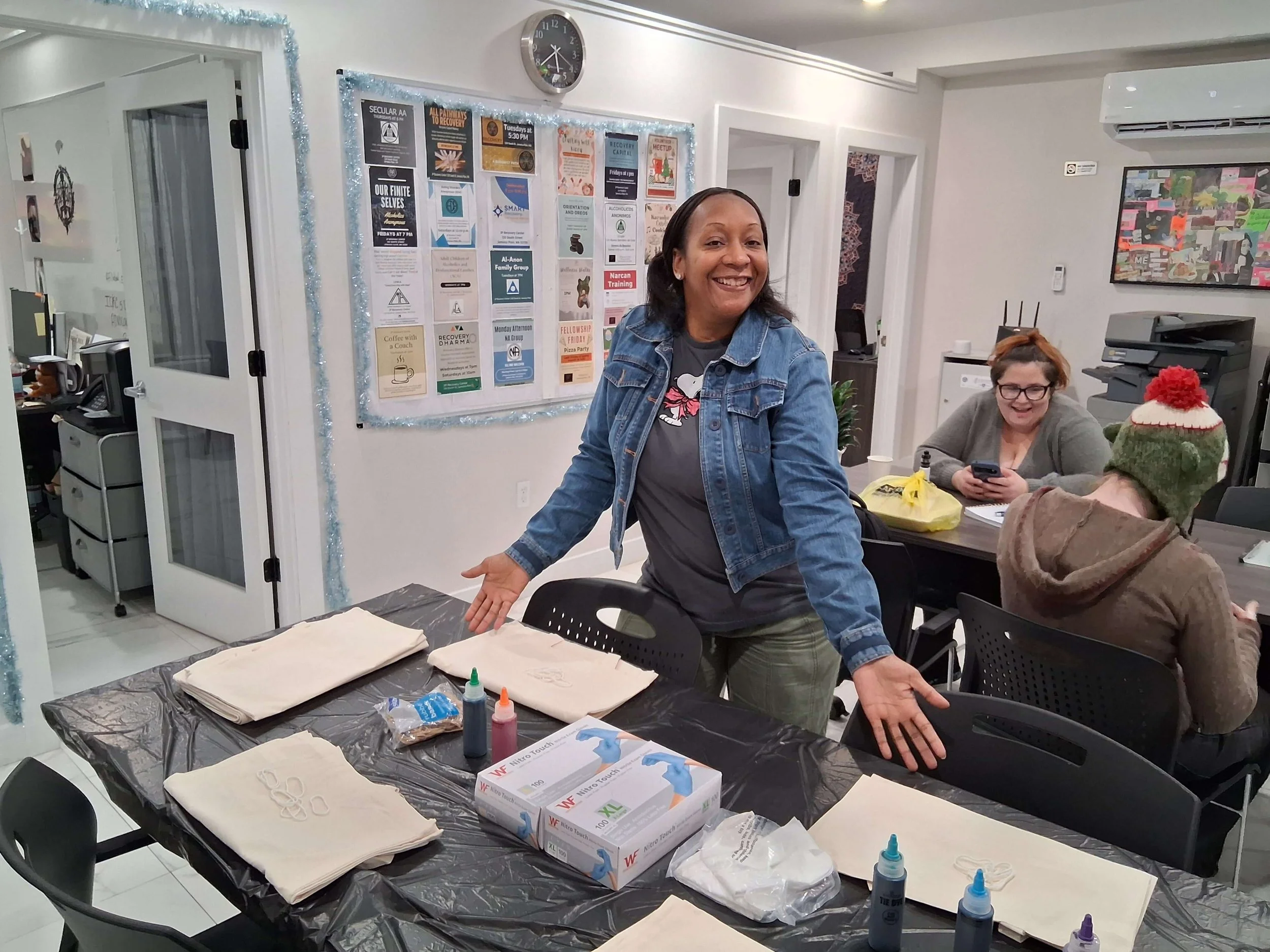 A woman wearing a denim jacket smiling and gesturing toward a table with napkins, gloves, and hand sanitizer in a community room with two other women in the background.