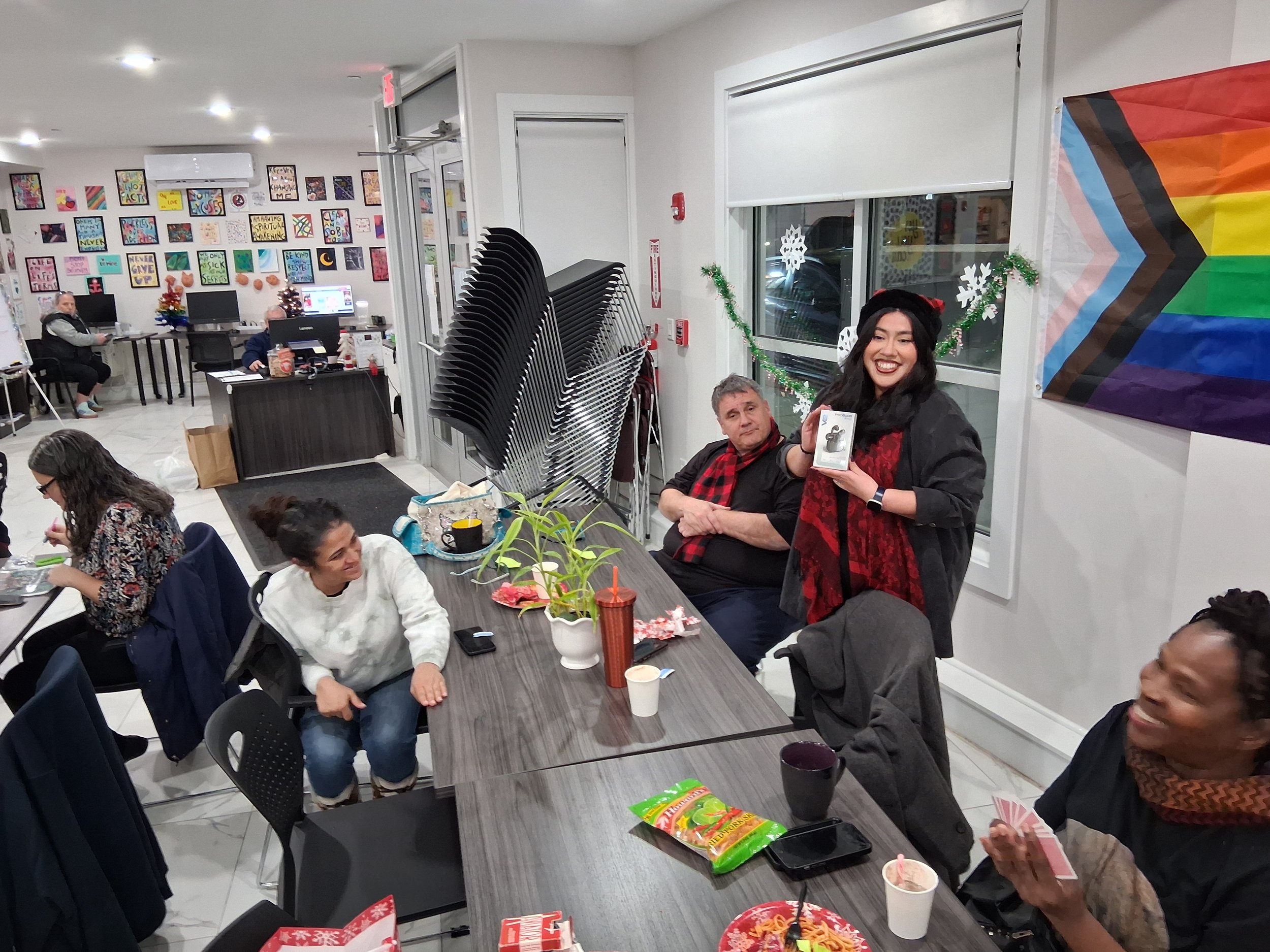 A group of people celebrating in a decorated room, with one woman standing and holding a gift box, smiling. The room has holiday decorations, including a Christmas tree, snowflake decorations, and a rainbow pride flag on the wall.