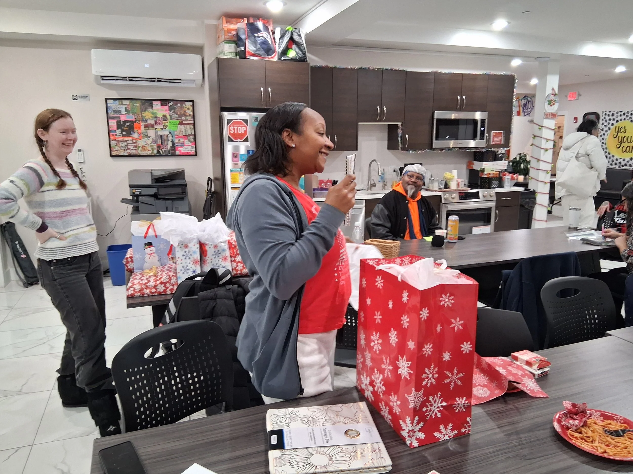 A group of people celebrating in a kitchen or dining area with Christmas decorations. A woman in red and gray is opening a gift, smiling. Other people are seated or standing around, and there are wrapped presents on the table.