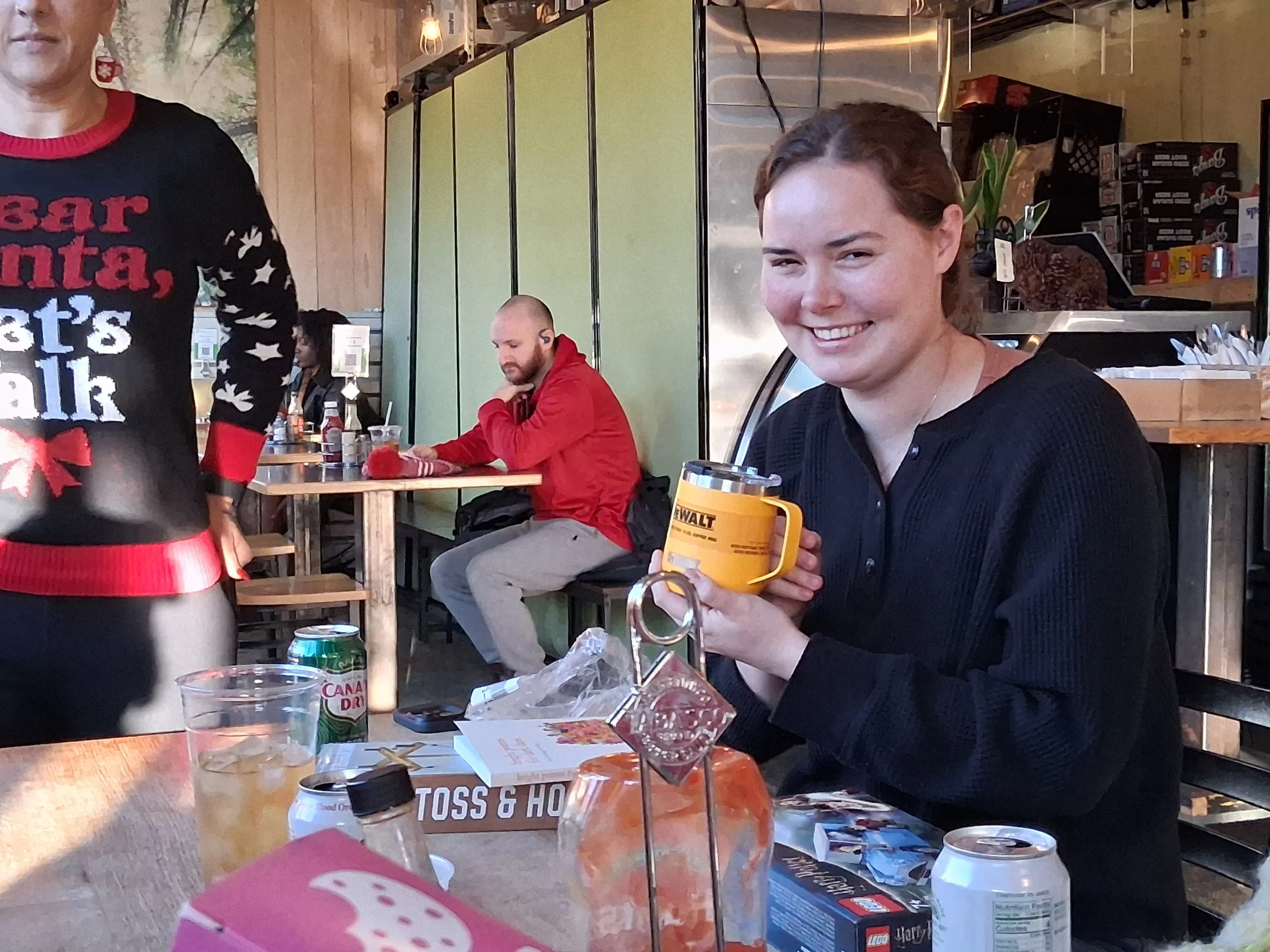 A young woman sitting at a table, smiling, and holding a yellow mug. On the table are various drink cans and food items. In the background, a man in a red hoodie is sitting at another table, using a mobile device, with a woman in a black shirt seated