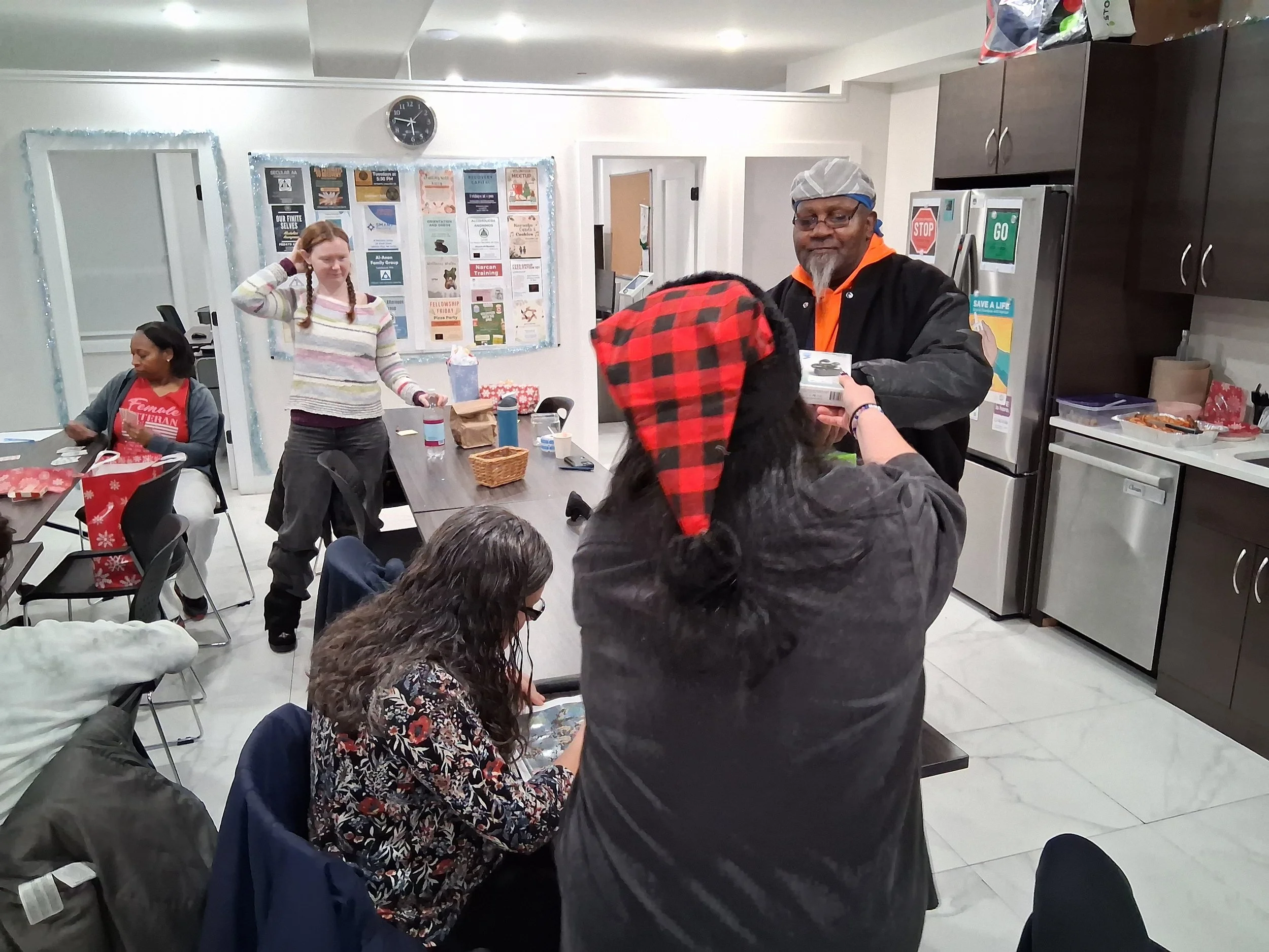 People gathered in a room with holiday decorations, one person is receiving a gift, and others are seated and standing around a table.
