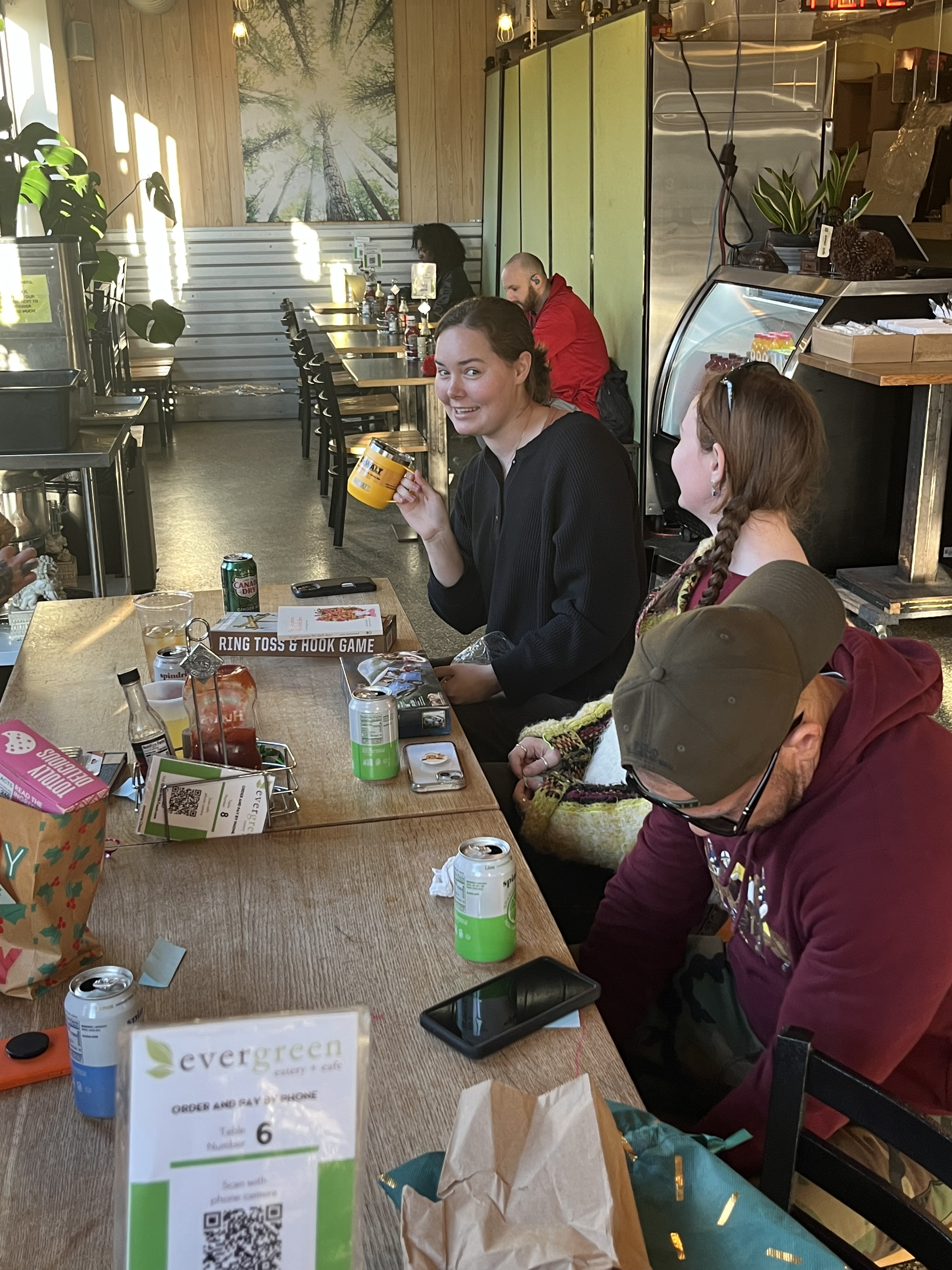 People sitting at a table in a cafe, with one woman smiling and holding a yellow mug. The table has drinks, a TV remote, and various items. A man in a maroon hoodie appears to be looking down, and other customers are seated in the background. There i