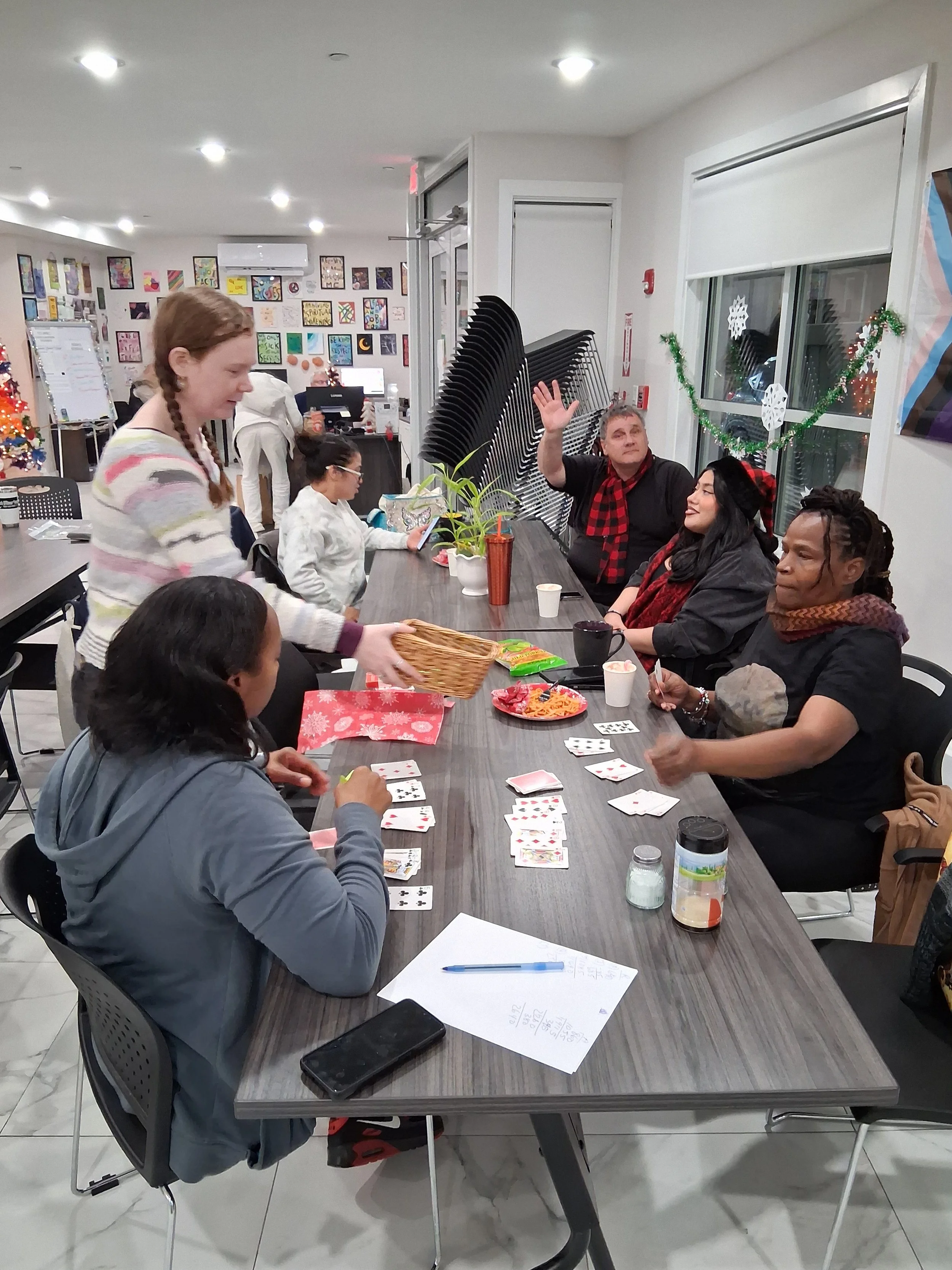 People gathered around a table playing cards, with a woman serving snacks. The room is decorated for Christmas with a small tree and holiday decorations.