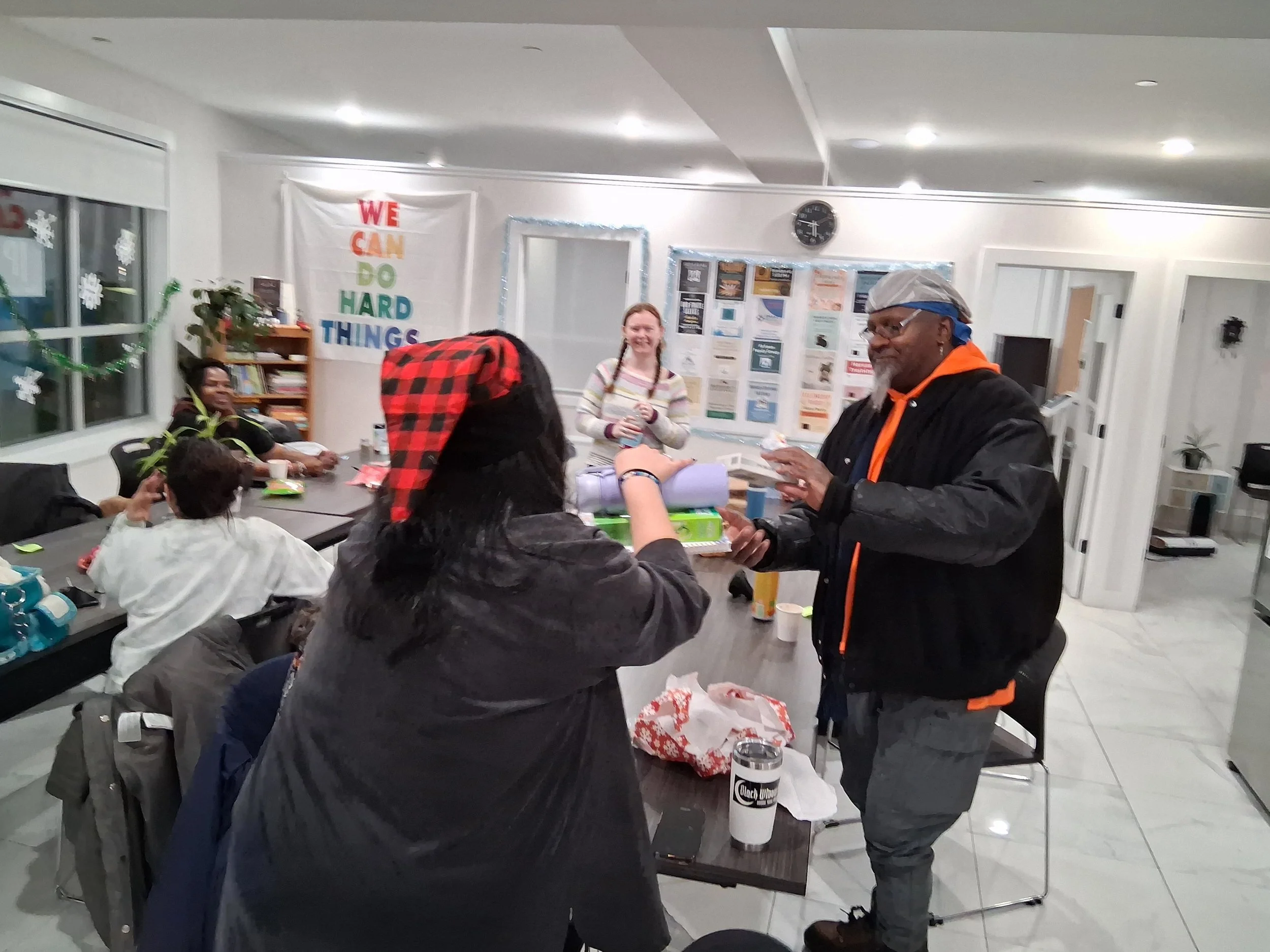 A man is giving a gift to a woman in a community room with several seated people. The room has festive decorations, a bookshelf, a clock, and a colorful banner that reads 'WE CAN DO HARD THINGS'.