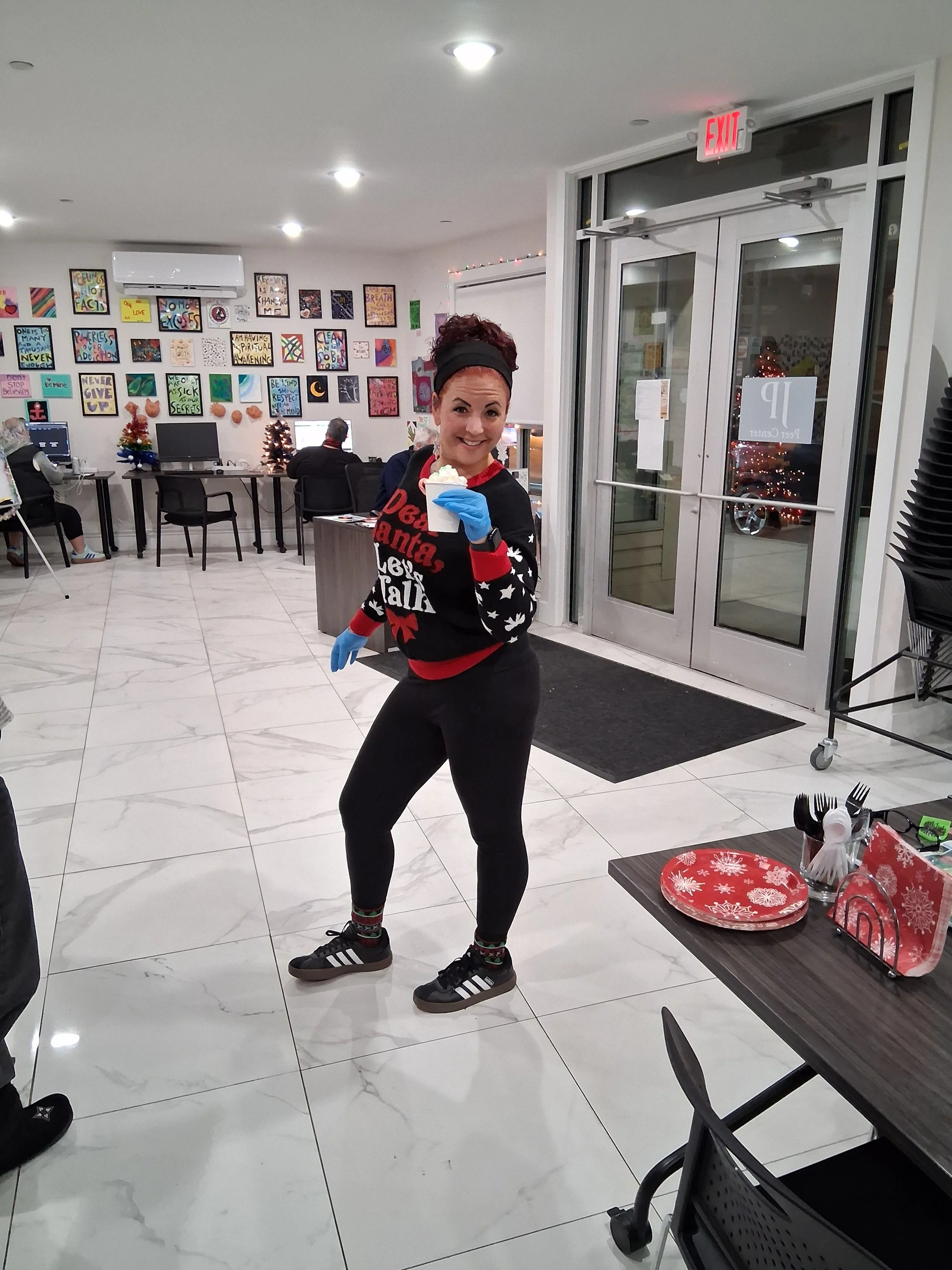 A woman smiling and holding a spoon with a dessert in a cup in a decorated indoor setting with artwork on the wall and Christmas decorations.