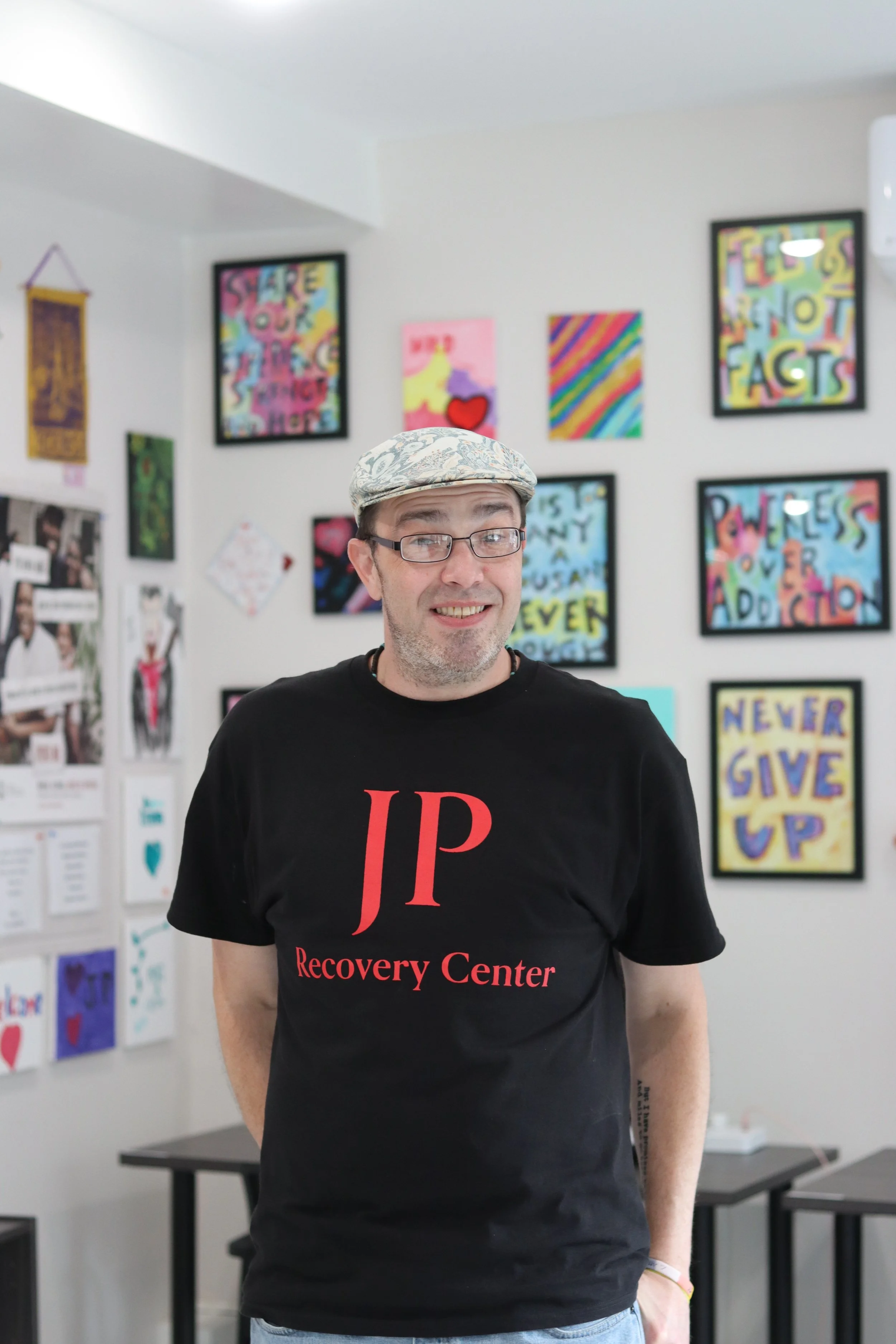A man wearing glasses, a patterned flat cap, and a black JP Recovery Center t-shirt, standing in an art-filled room with colorful paintings and posters on the walls.