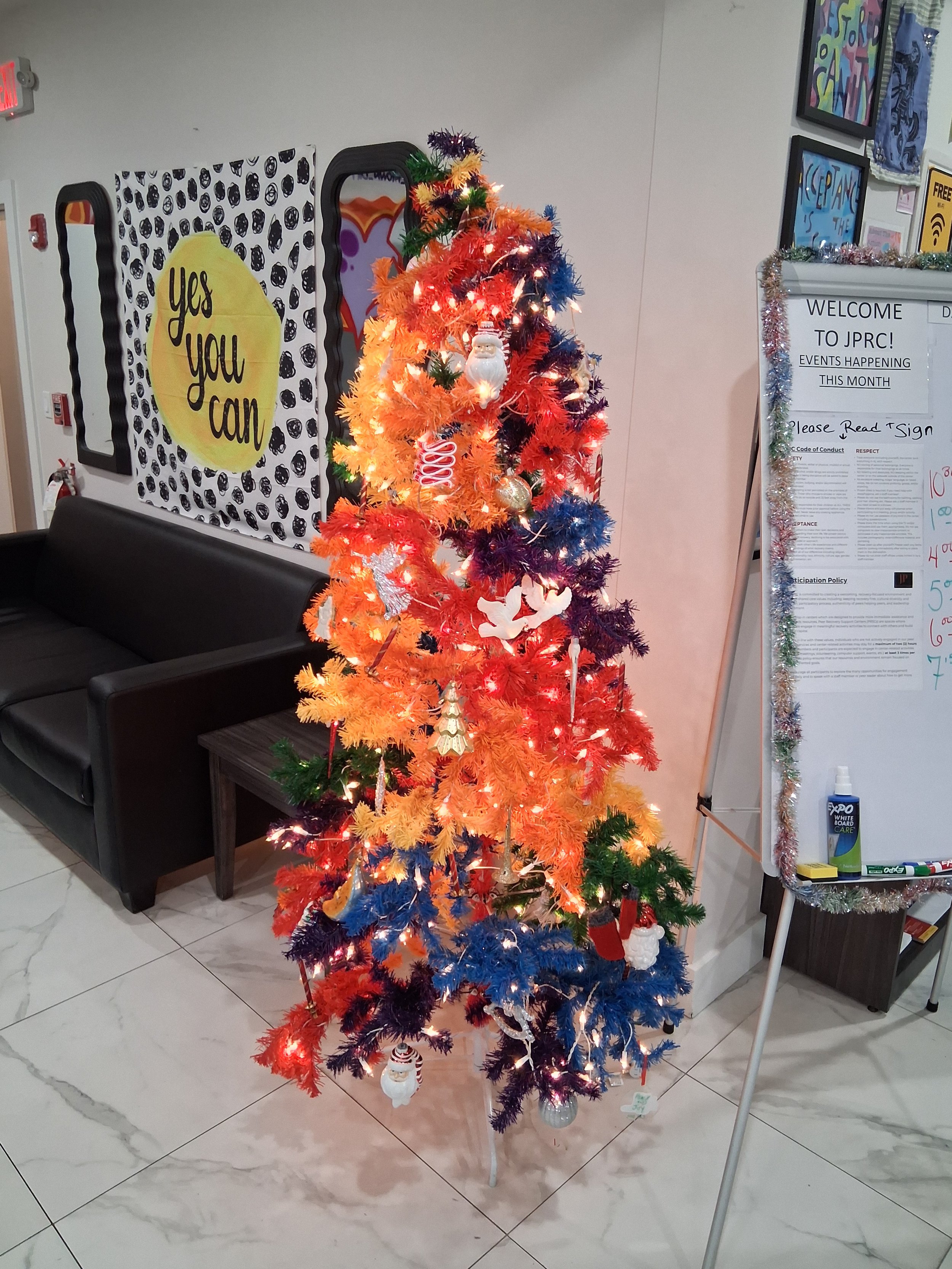 Colorful artificial Christmas tree decorated with various ornaments and lights in a room with white walls, black couch, and whiteboard.