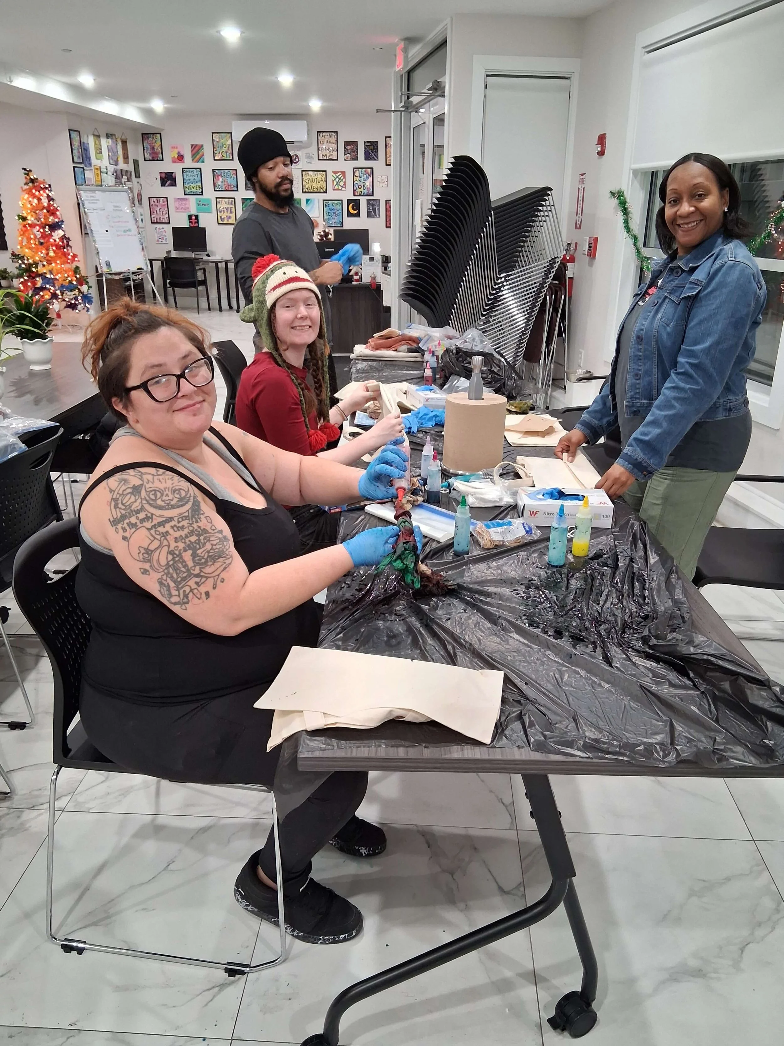 Four people sit at a table covered with black plastic, engaging in a tattoo or art activity. One woman with glasses and a tattoo on her arm smiles at the camera, while another woman in a red hat looks at her. A man stands in the background with glove