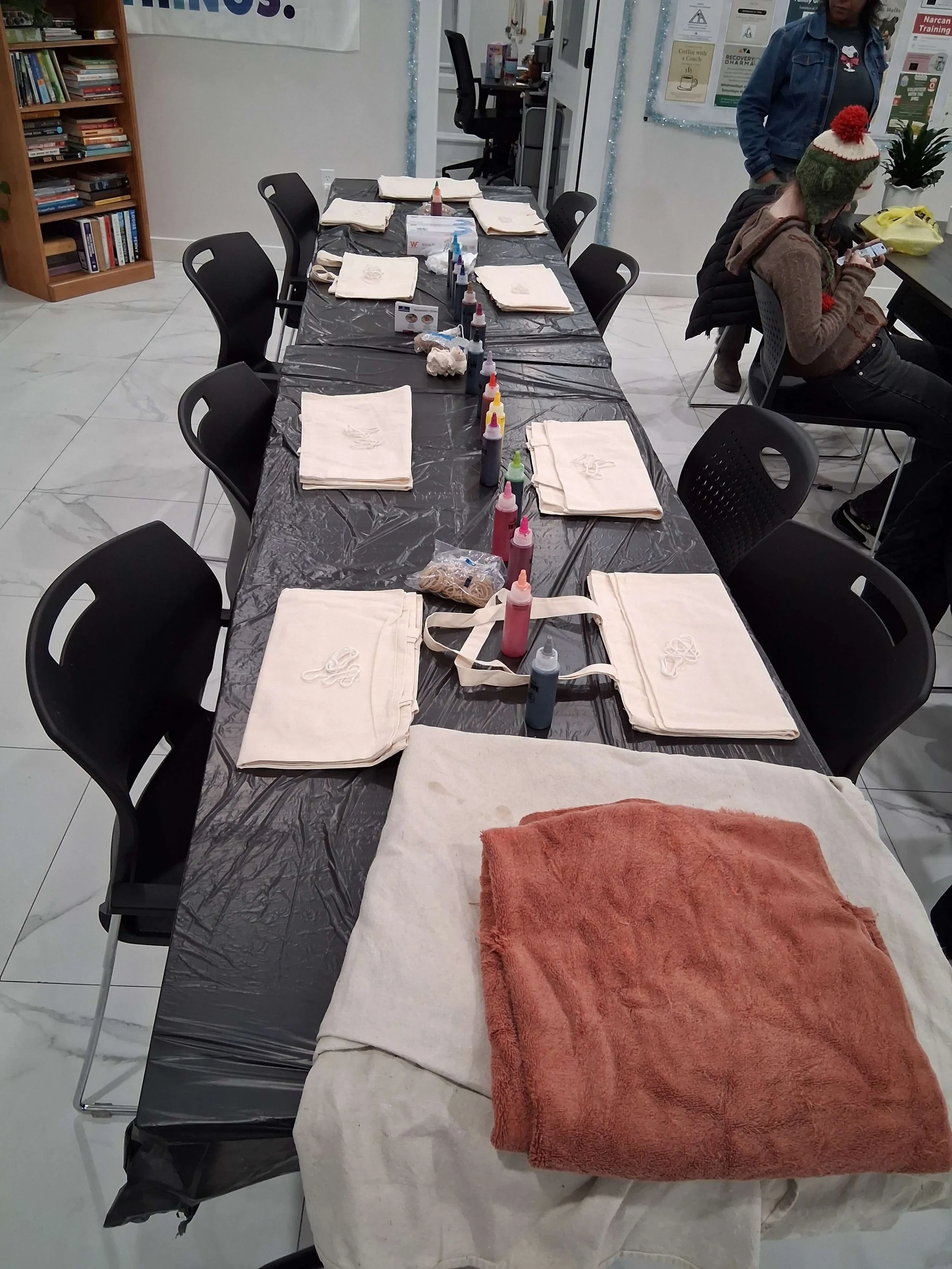 A long table covered with a black plastic tablecloth, set up for a crafting activity with paper bags and bottles of paint, surrounded by black chairs. There is a folded towel on a white cloth at one end of the table. In the background, two people are