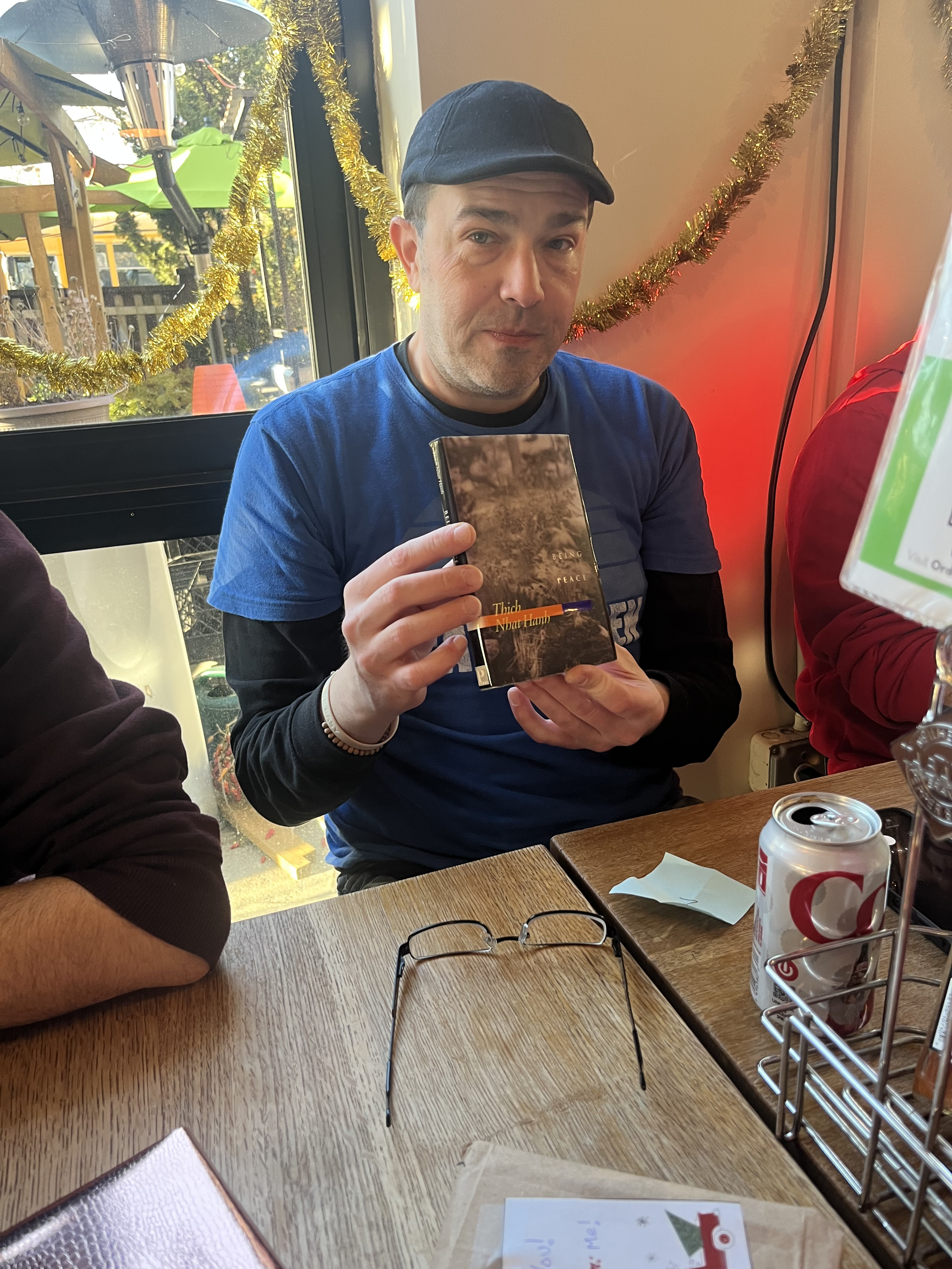 Man wearing a black cap, blue T-shirt, and black long-sleeve shirt holding a book and sitting at a table in a decorated restaurant.