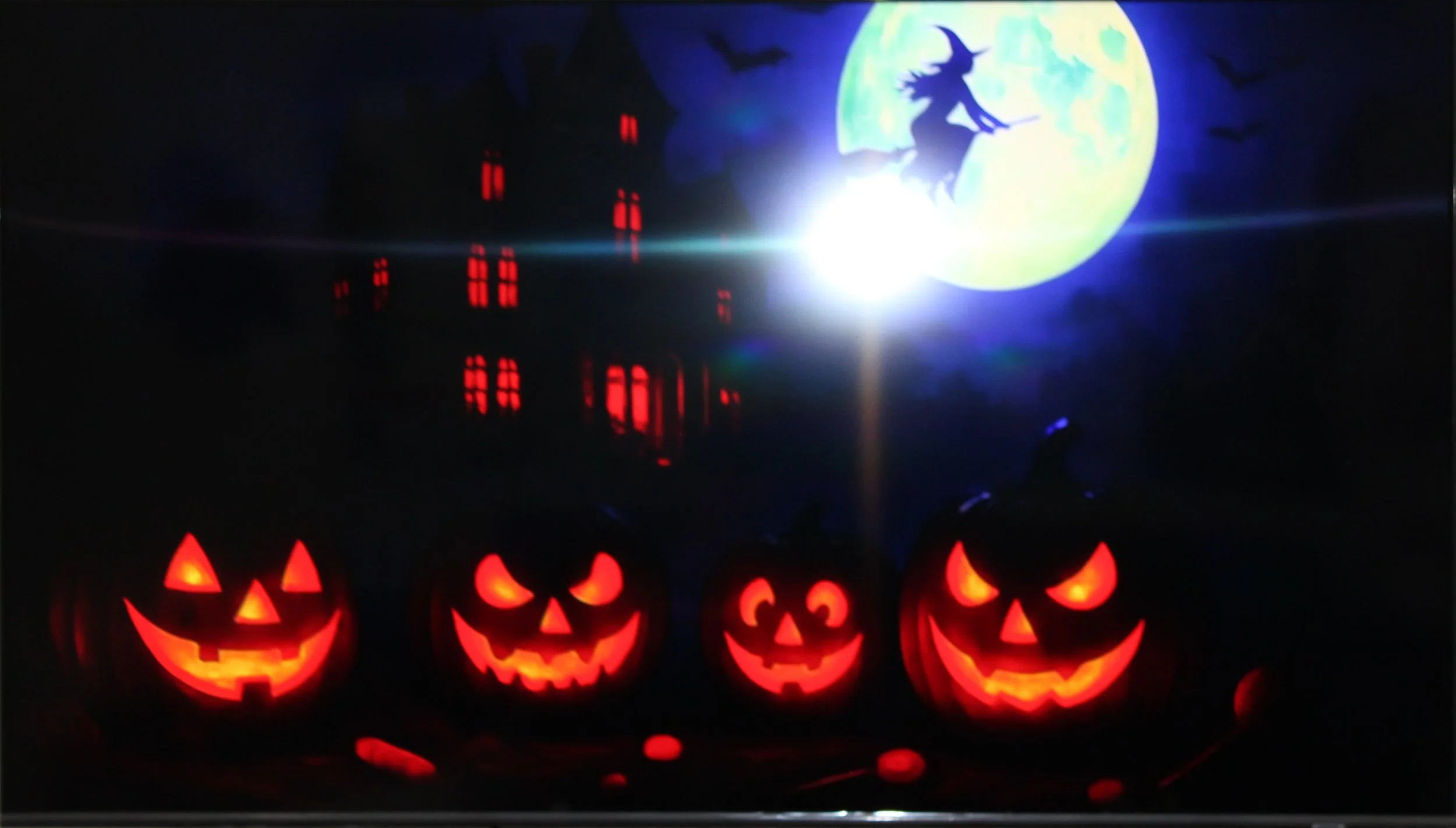 Four illuminated Halloween pumpkins with carved faces in front of a dark background, with a large full moon featuring a silhouette of a witch on a broomstick and a haunted house with red lit windows under a night sky.