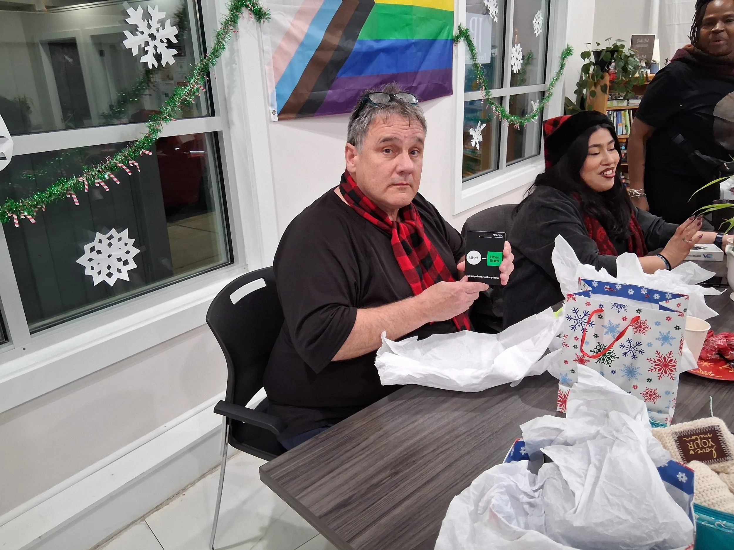 A man with glasses on his head and a red and black checkered scarf around his neck sitting at a table with Christmas decorations in the background, holding a phone displaying the Uber app. Beside him is a woman smiling, with gift wrapping and Christm