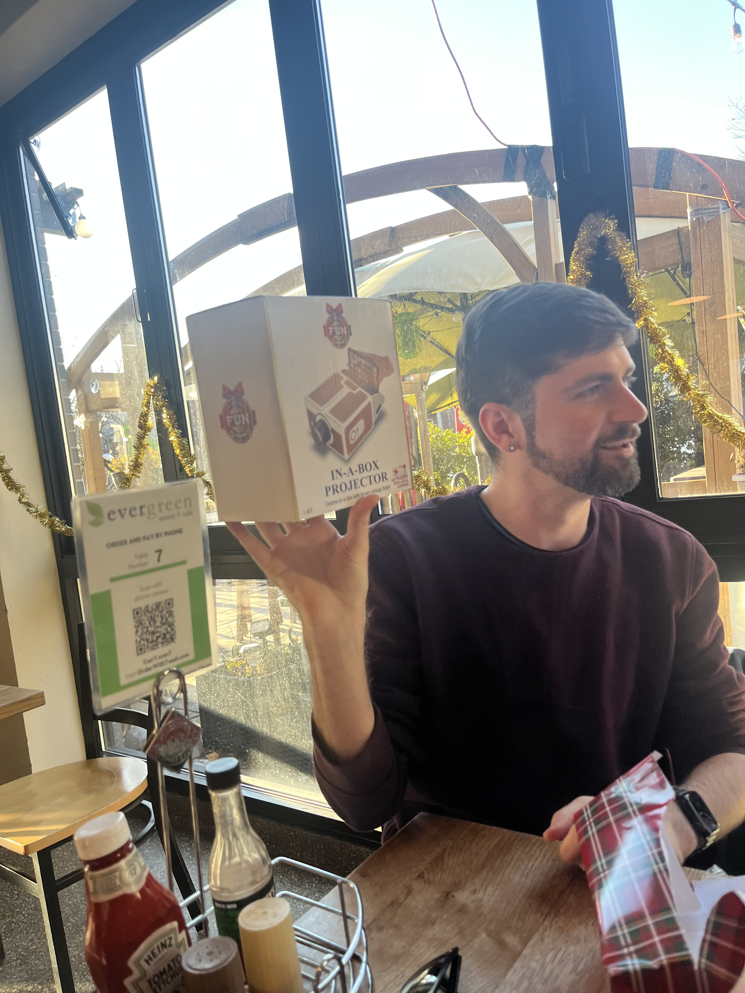 Man sitting at a restaurant table holding a boxed in-a-box projector. Christmas decorations on the window behind him.