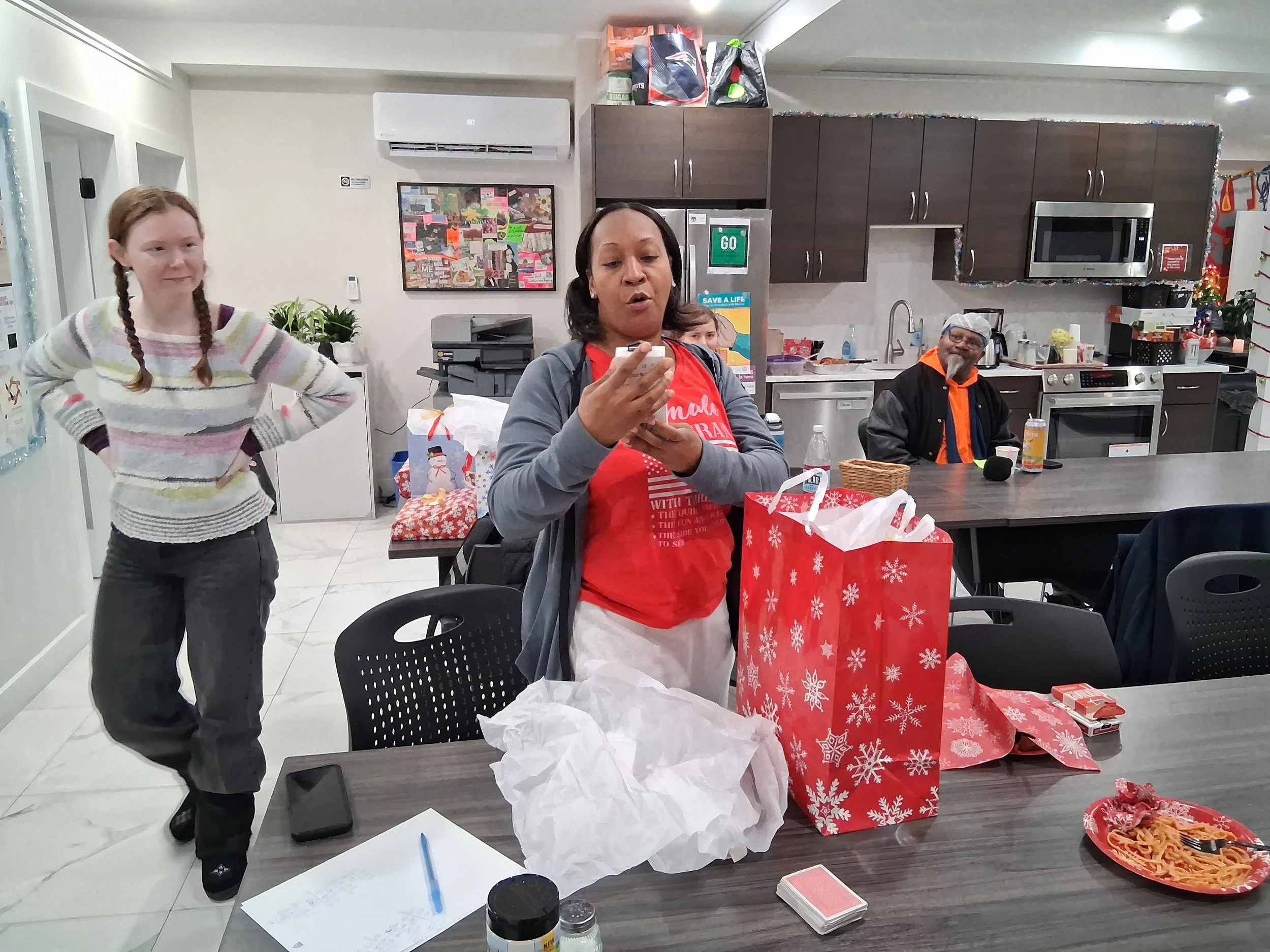 People gathered in a kitchen with Christmas decorations, woman in red shirt opening a gift, girl in striped sweater standing with hands on hips, man smiling at the back, holiday-themed gift bags and a plate of spaghetti on the table.
