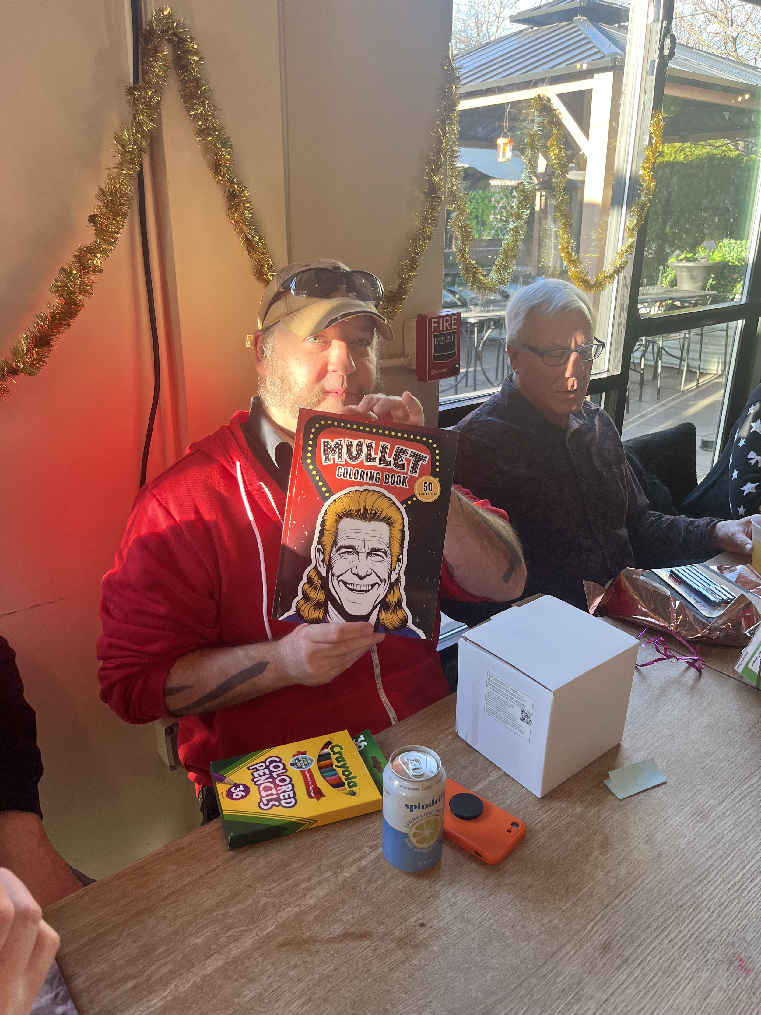 A man in a red jacket holding a 'Mullet Coloring Book' at a table during a gathering, with wrapped gifts, a soda can, and Crayola colored pencils on the table, in a room decorated with gold tinsel and large windows showing an outdoor patio.