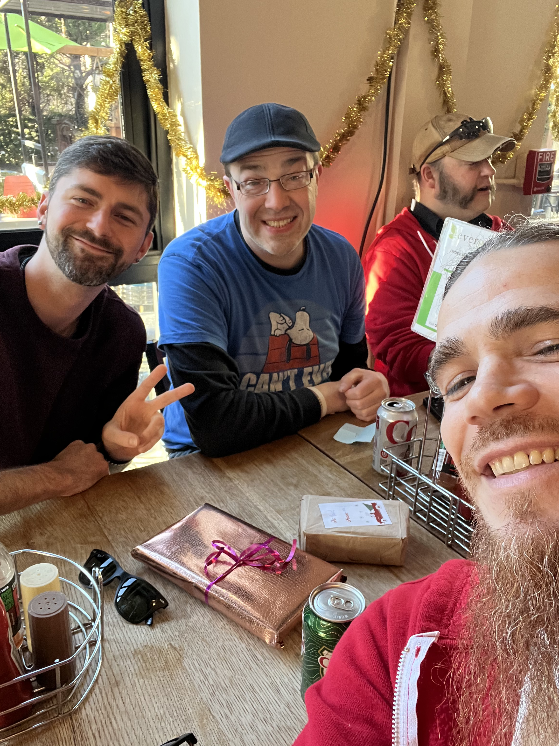 Four men sitting at a wooden table in a festive setting, smiling at the camera. The man in the foreground has a beard and is taking a selfie. The others are sitting side by side, with one making a peace sign. There are wrapped gifts, cans, and sungla
