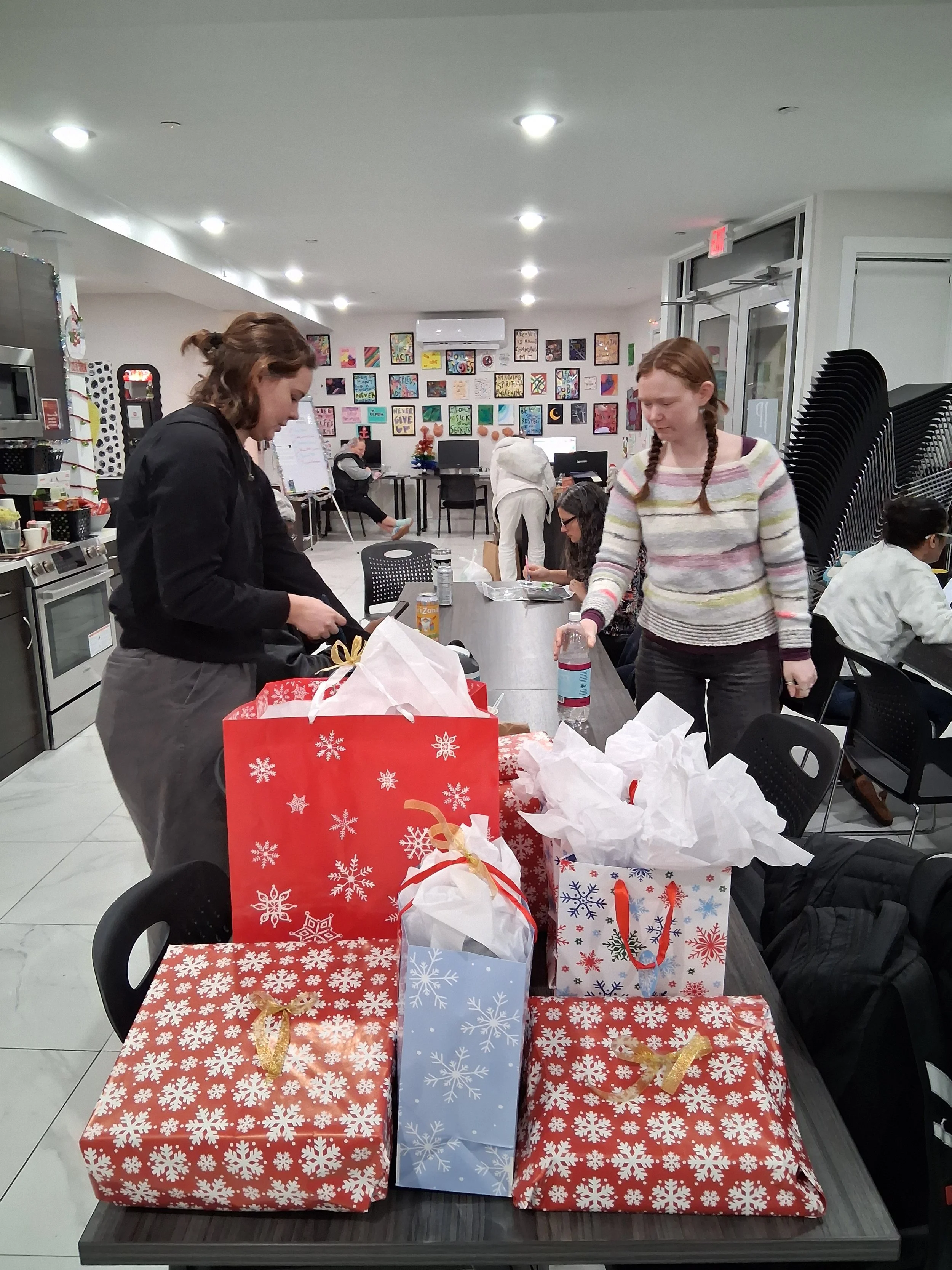 Table with Christmas presents wrapped in red, white, and blue paper with snowflake designs, some with ribbons, in a room decorated with colorful artwork and Christmas decorations.