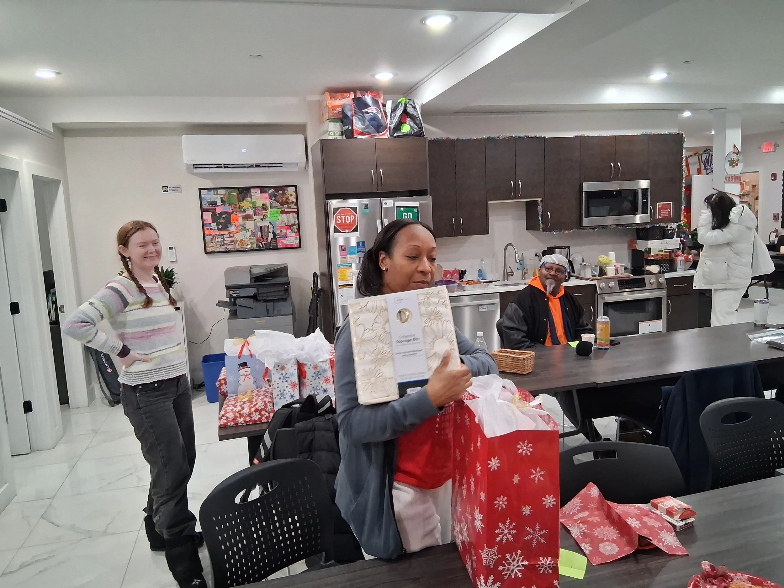 People gathered in a kitchen-like room with wrapped holiday gifts on the table, celebrating Christmas with festive decorations.