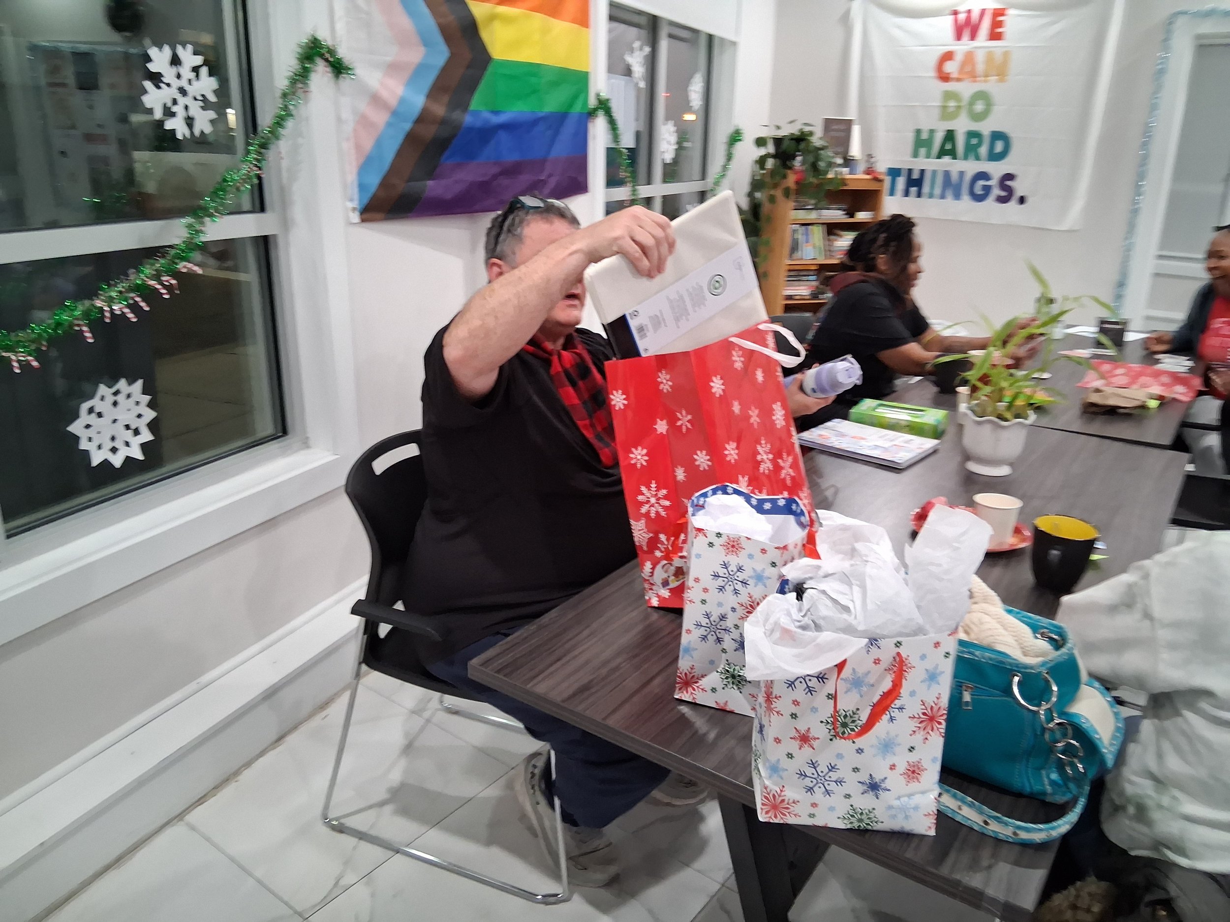 Person opening a gift bag at a holiday gathering with others sitting around the table, decorated with Christmas-themed bags, papers, and cups, in a room with festive decorations including snowflakes and a rainbow pride flag.
