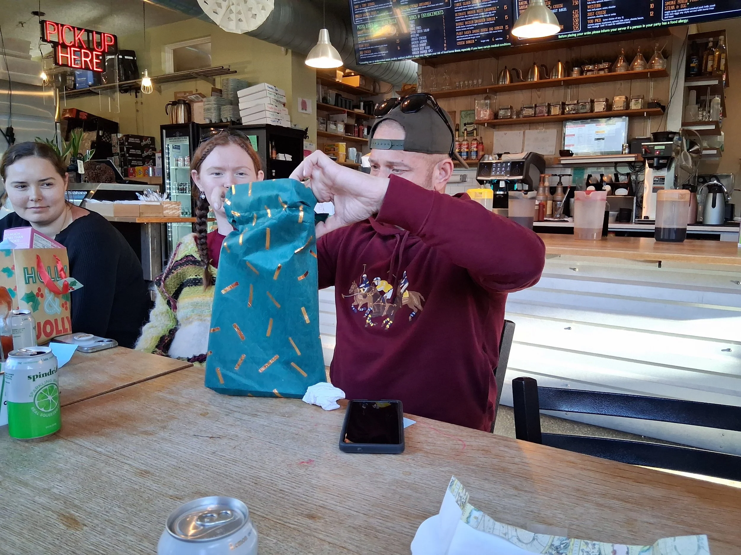 A man wearing a burgundy hoodie with a print of medieval knights and a backward baseball cap is opening a gift bag at a restaurant or cafe with two women sitting nearby. One woman has braided hair and is wearing a multicolored sweater, the other has 