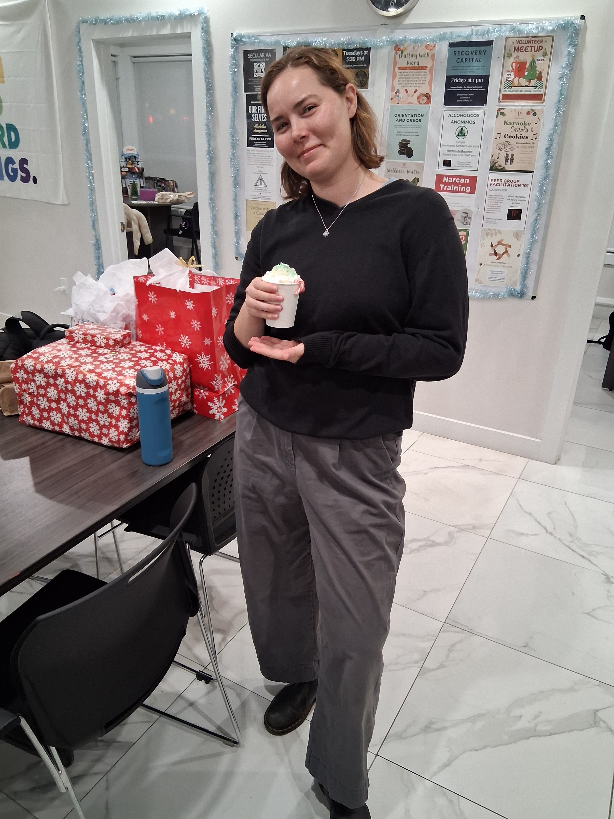 A woman with shoulder-length brown hair wearing a black sweater and gray pants, standing indoors while holding a cup of ice cream.