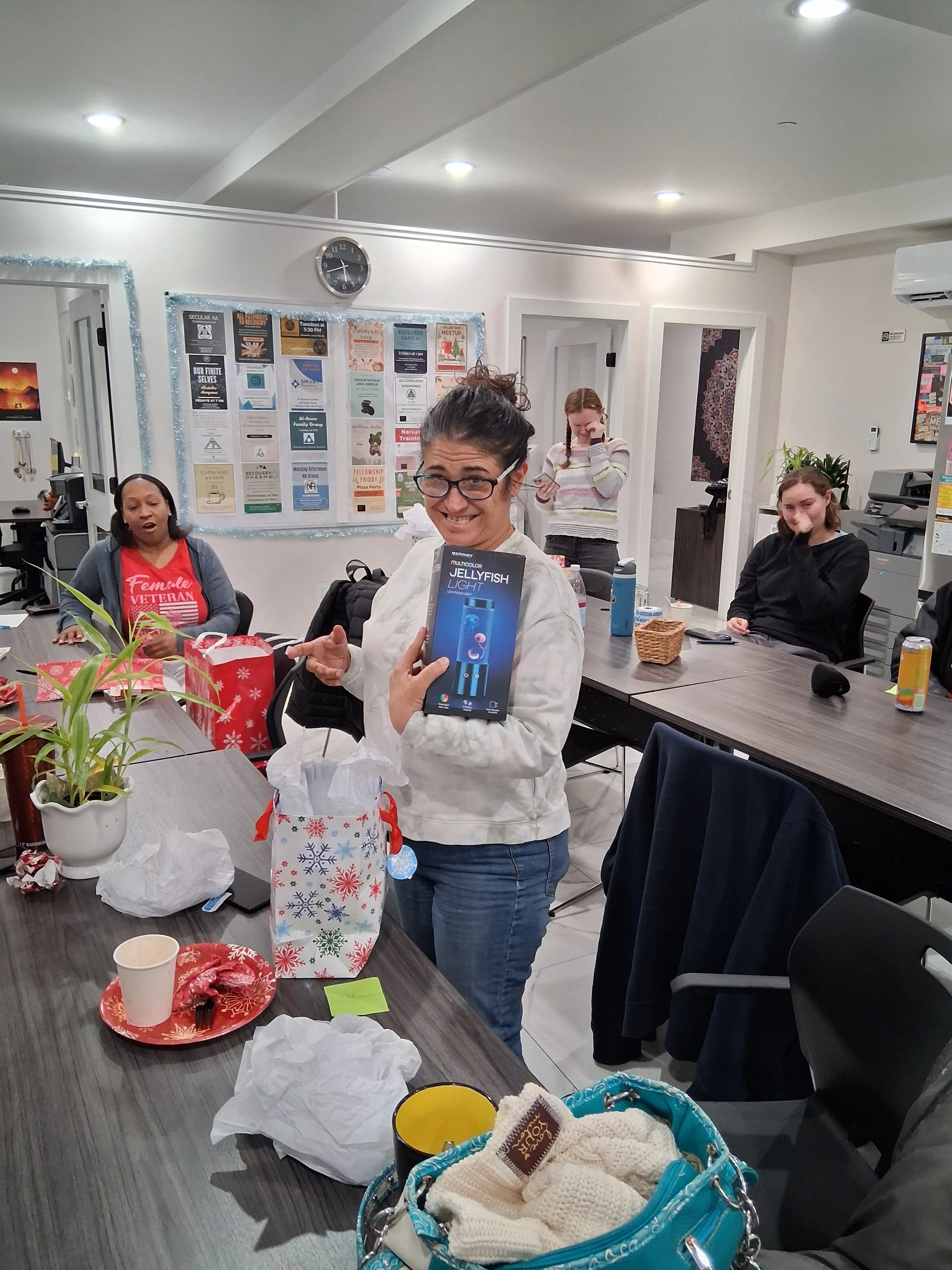 A woman holding a box of Jellyfish Light while smiling at a gathering around a table, with other women seated and standing, in a room with holiday decorations and gifts.