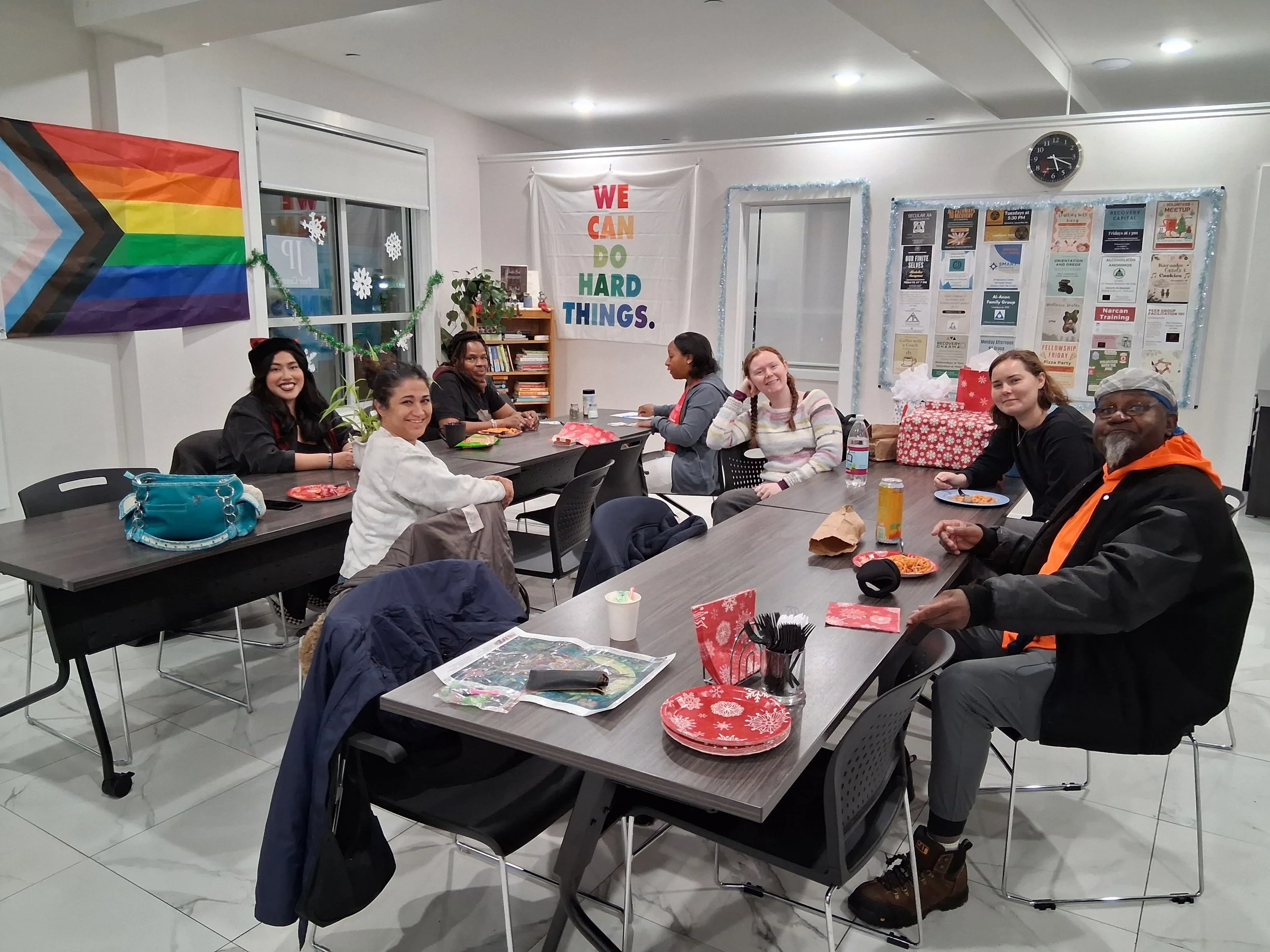 A diverse group of seven people sitting around tables in a room decorated for the holidays with a large rainbow pride flag, a banner reading 'WE CAN DO HARD THINGS,' and holiday-themed paper plates, napkins, and gift boxes, enjoying snacks and drinks