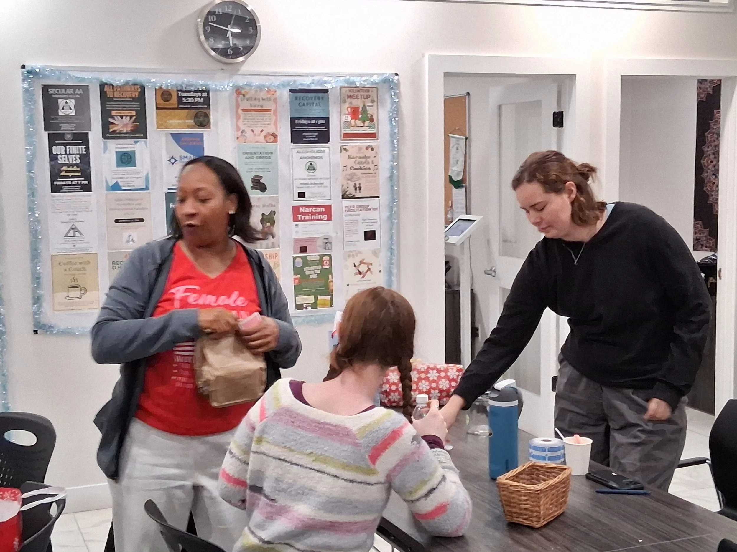 Three women in a room with a table, two are standing and one is sitting. The woman on the left is holding a paper bag, the woman on the right is reaching across the table, and the woman sitting has red hair and is wearing a striped sweater. There are