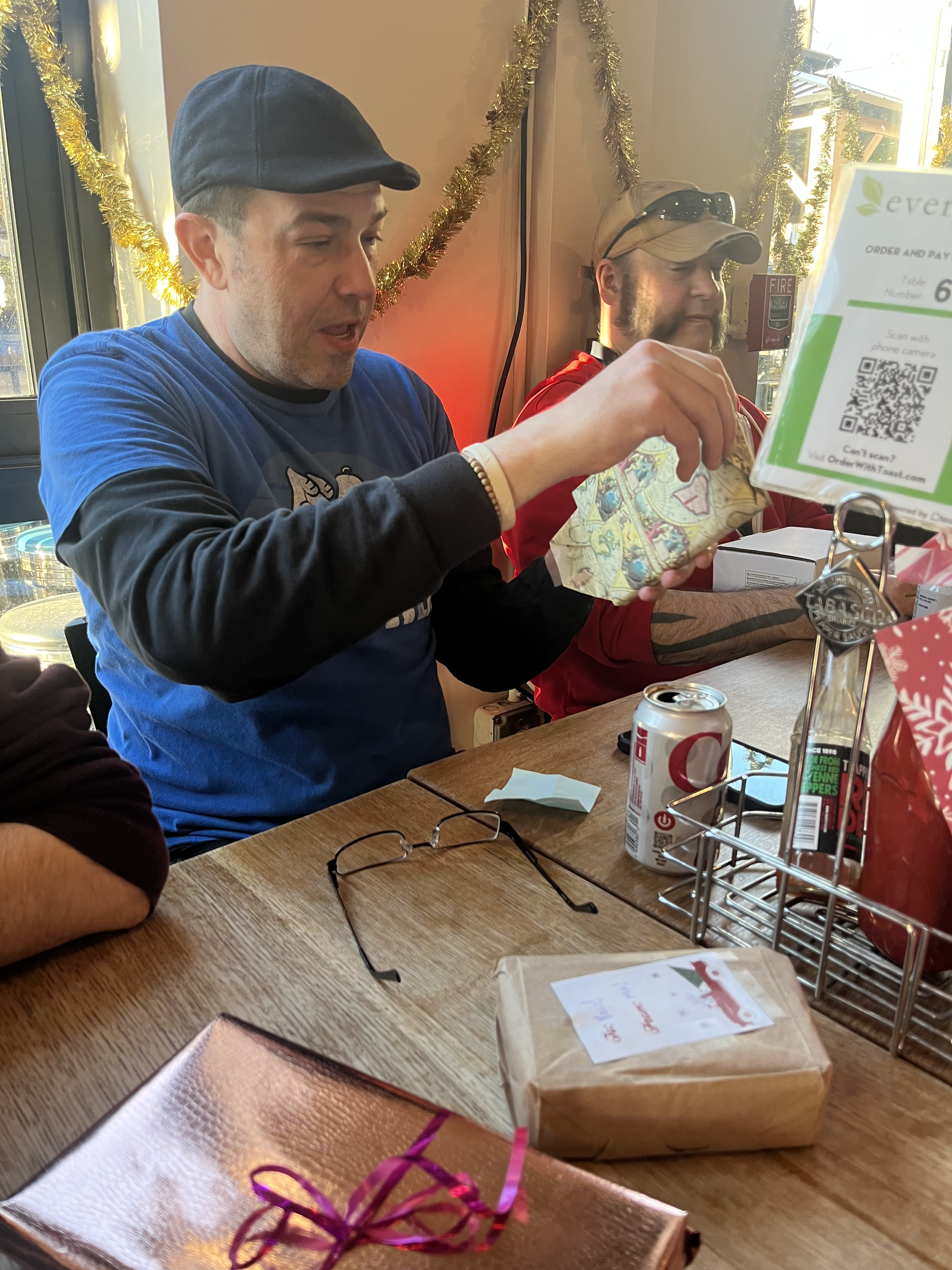 Two men sitting at a table opening a wrapped gift during a holiday celebration. Decorations and a gift bag are visible on the table.