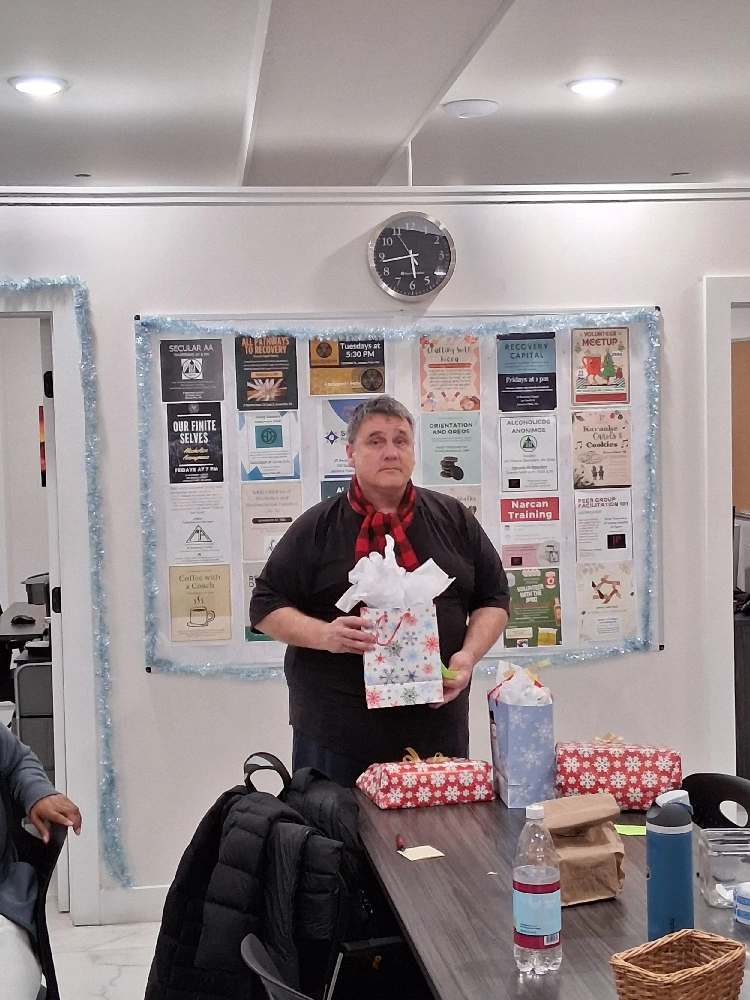 A man standing in front of a decorated bulletin board holding a gift bag, wearing a red and black plaid scarf. The table in front of him has wrapped presents, a water bottle, and other items. The bulletin board behind him has various posters and noti