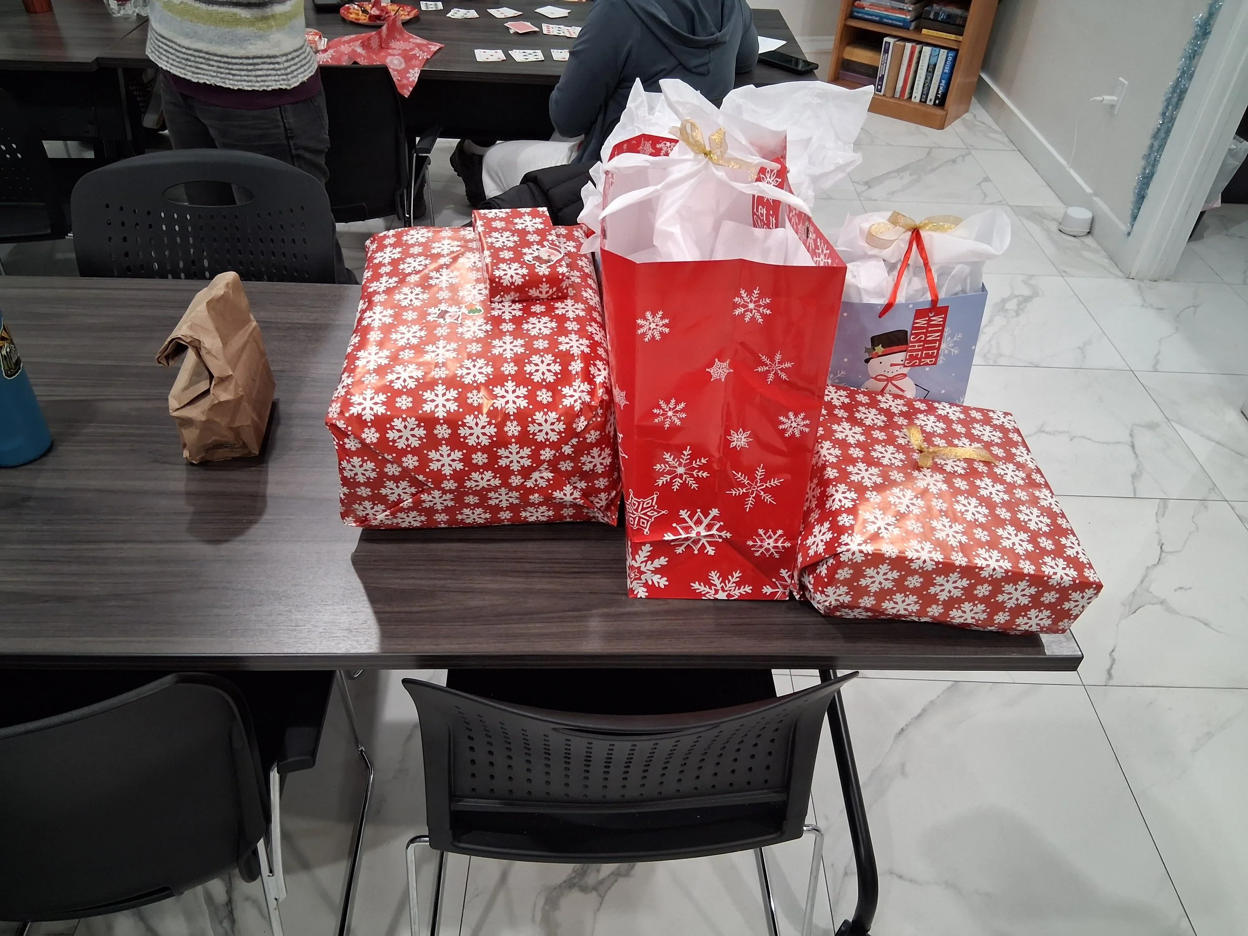A dark wooden table with four wrapped presents in red and white snowflake designs and white tissue paper on top, surrounded by black chairs, in a room with light-colored tiled floors, bookshelves, and people in the background.