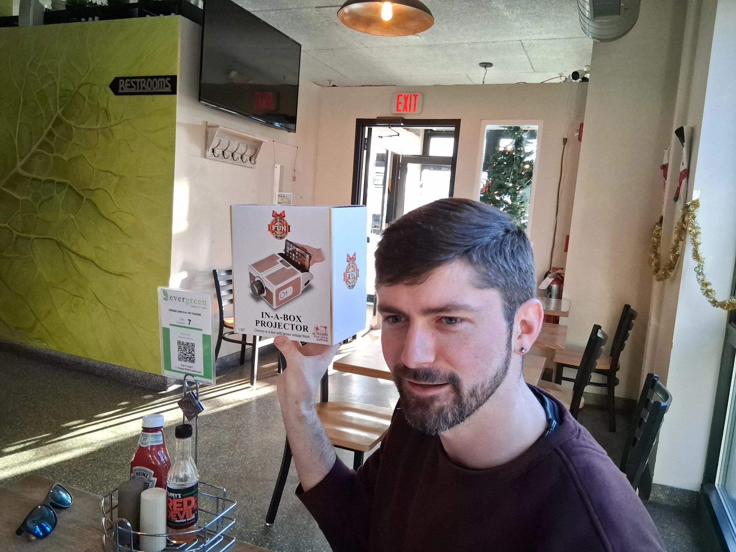 A man with short dark hair and a beard sitting in a cafe, holding a boxed 'In-A-Box Projector' toy. The cafe has holiday decorations, a Christmas tree visible through the window, and condiment bottles on the table.