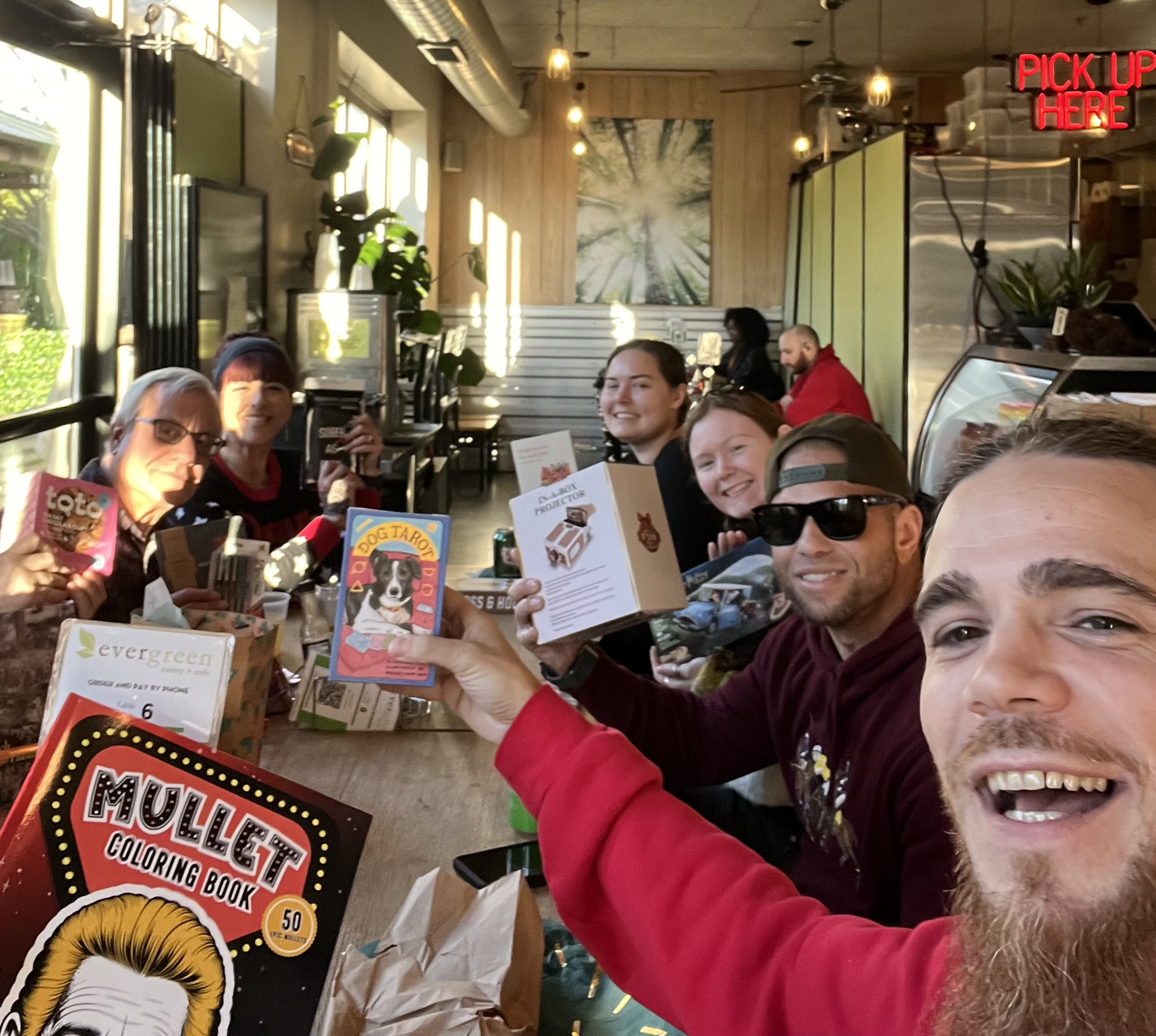A group of people sitting at a restaurant table, smiling and holding various game and book items, with sunlight coming through the windows and a neon sign that says 'PICK UP HERE' in red.