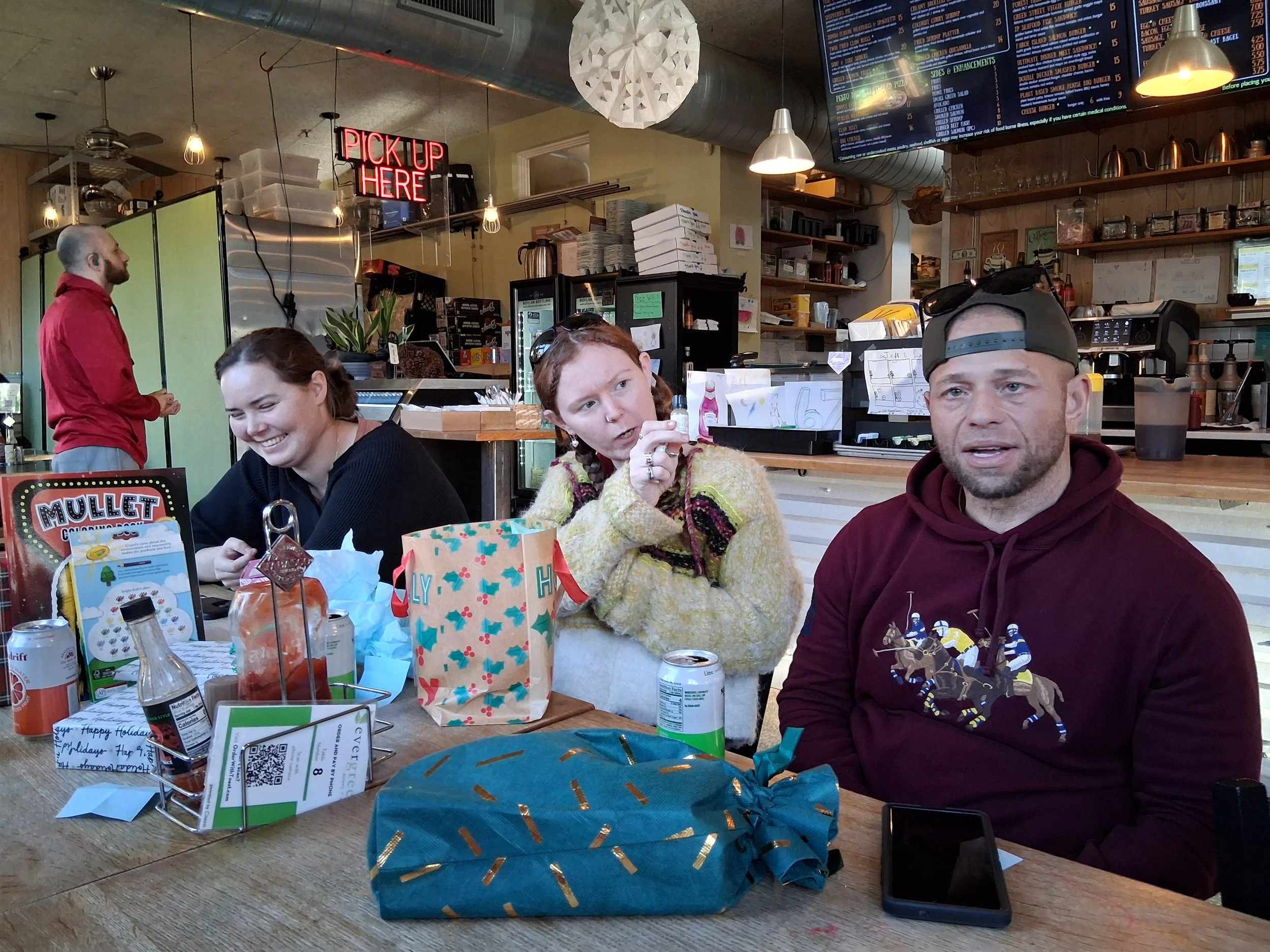 Four people sit at a wooden table in a casual restaurant or cafe, with various items and beverages in front of them. A woman in black is laughing, a young girl in a colorful sweater is listening, a man in a maroon hoodie with a illustrated sports sce