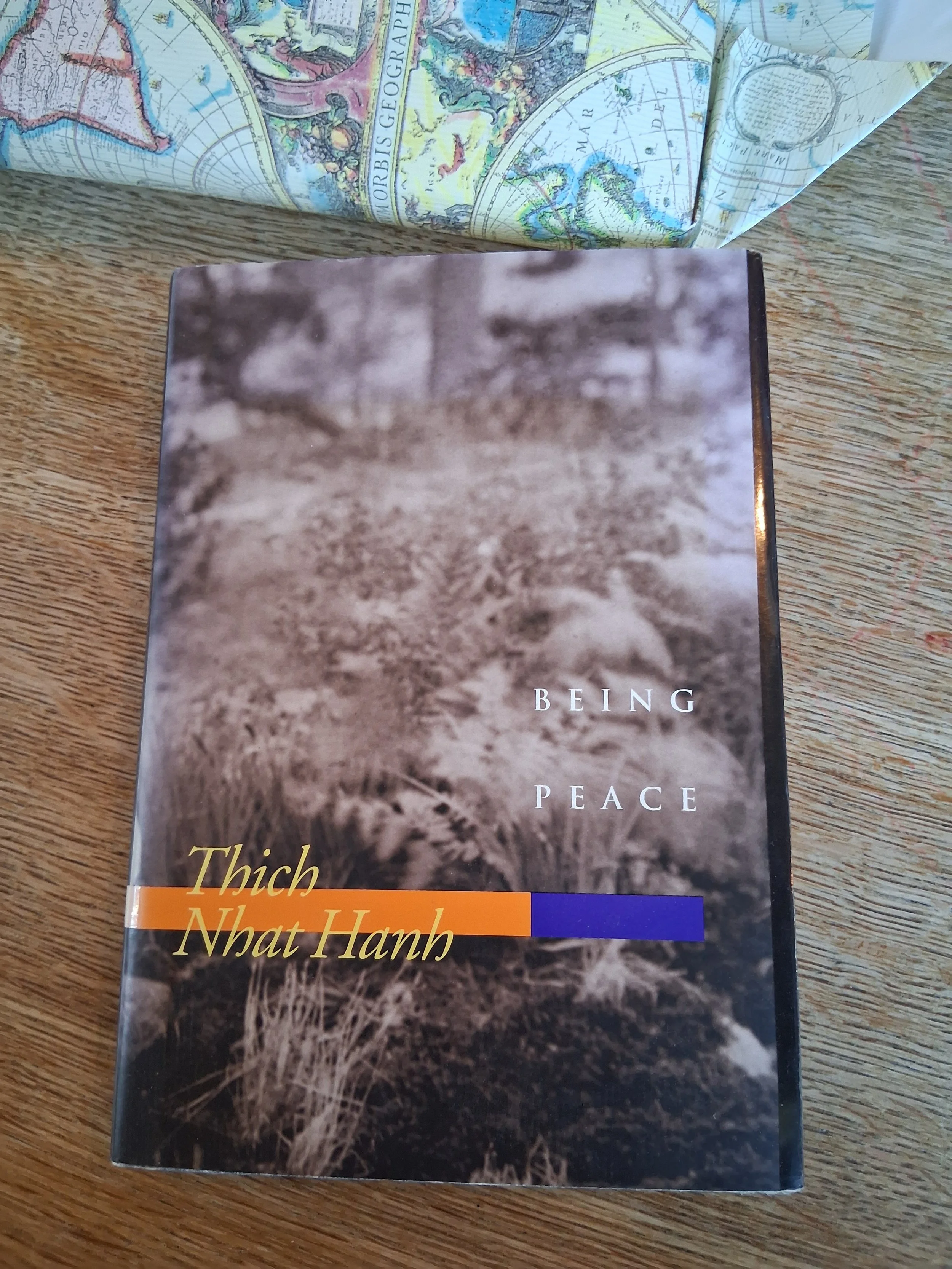 A book titled "Being Peace" by Thich Nhat Hanh on a wooden surface, with a partially visible map or illustrated paper in the background.