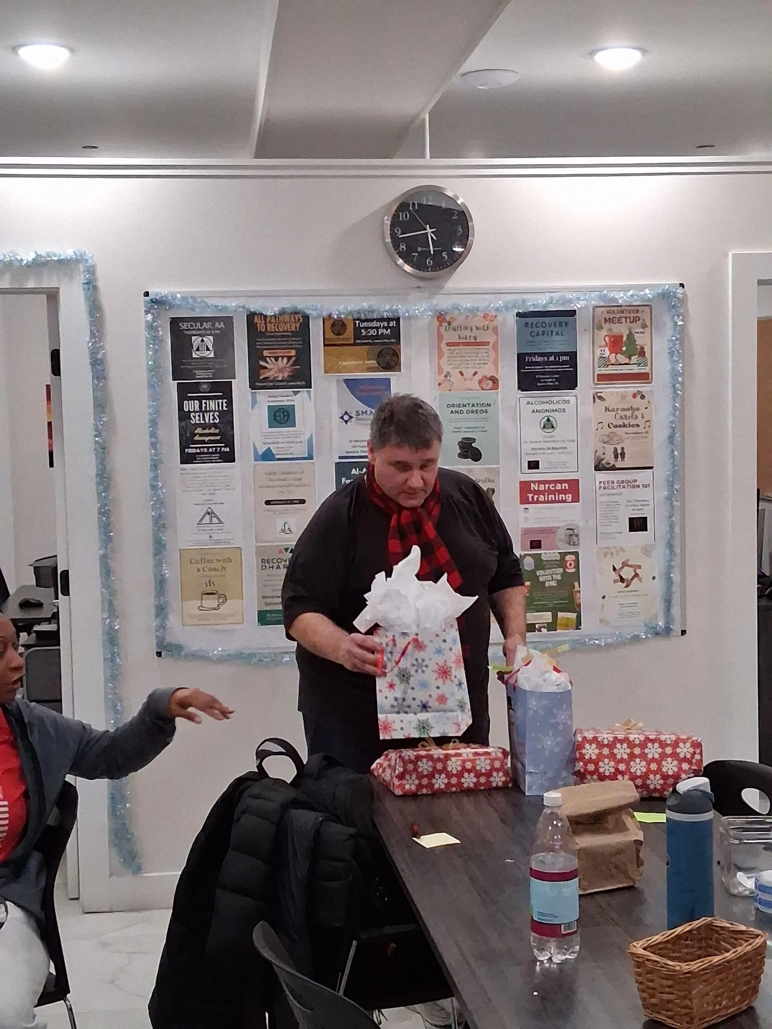 A man with short gray hair, wearing a black shirt and a red checkered scarf, opening gift bags at a holiday celebration in a room with a bulletin board and a clock on the wall.