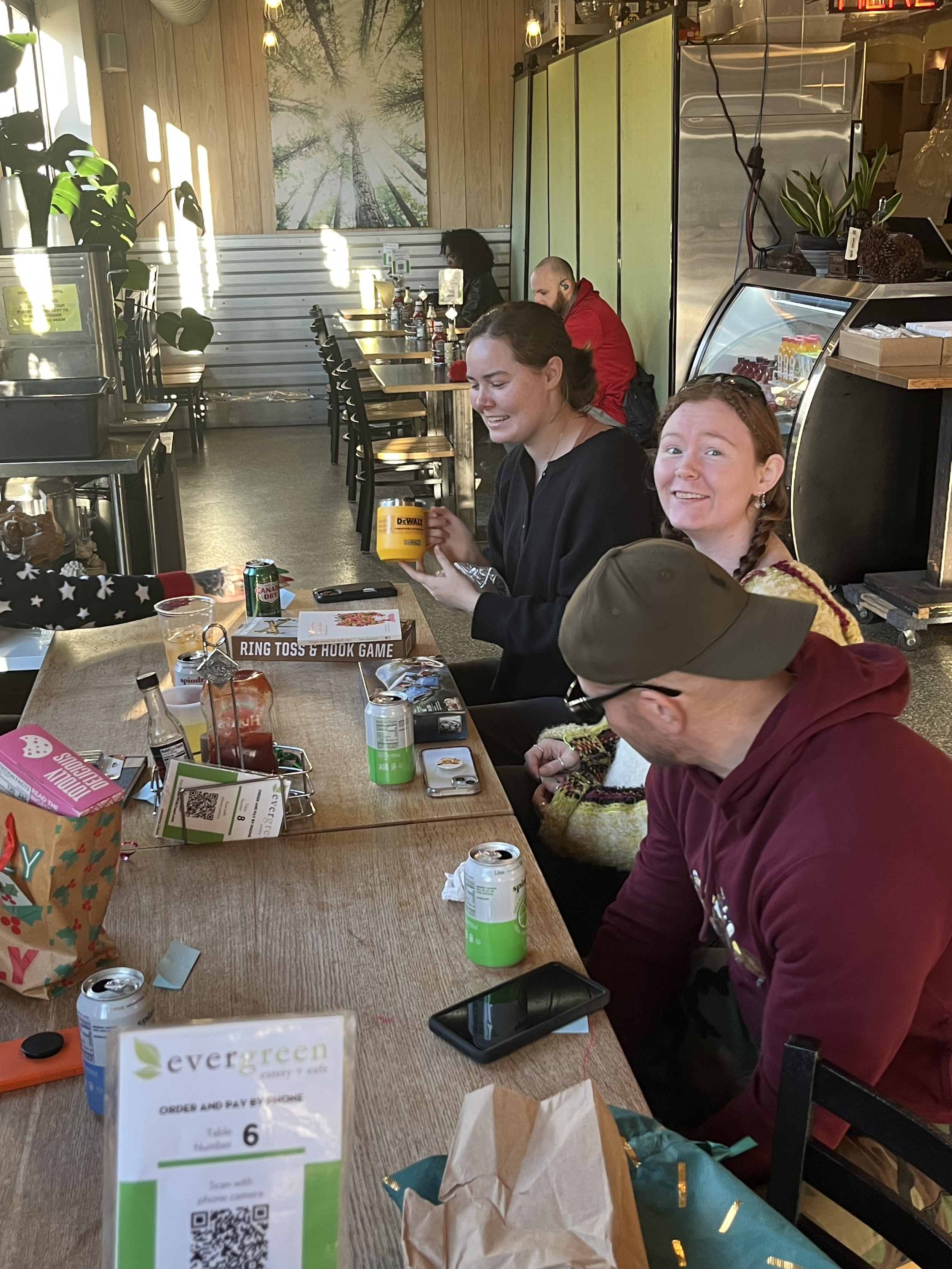 People sitting at a long wooden table in a cafe, with drinks and snacks. Two women smiling at the camera, one holding a yellow cup, and a man in a baseball cap looking down. In the background, a wall with a forest mural and a row of chairs and tables